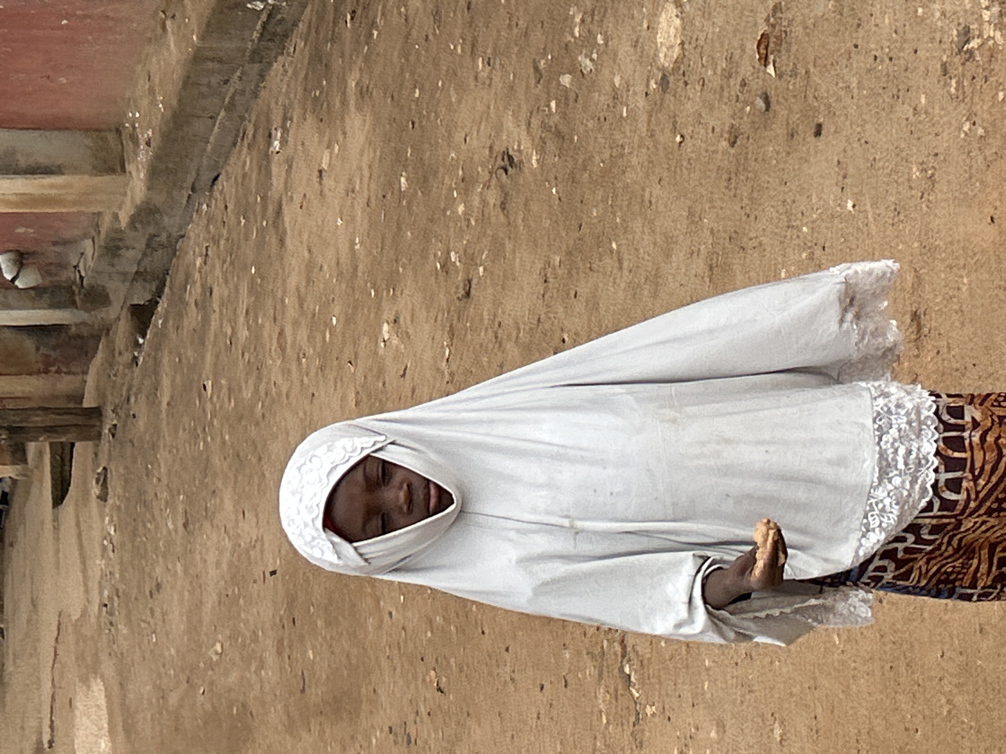A woman wearing a white headscarf and robe stands on a dirt ground surface with what appears to be a building to her left. The woman's head is tilted downward. She has dark skin and her eyes are closed. Her headscarf has white lace trim. She is wearing a patterned skirt with brown, black, and white colors. One of her feet is visible. The ground is textured and mottled with brown and gray. To the left, part of a building is visible with reddish tones and a structural element that could be a beam or ledge. The image appears to have been taken in Jos, Nigeria.