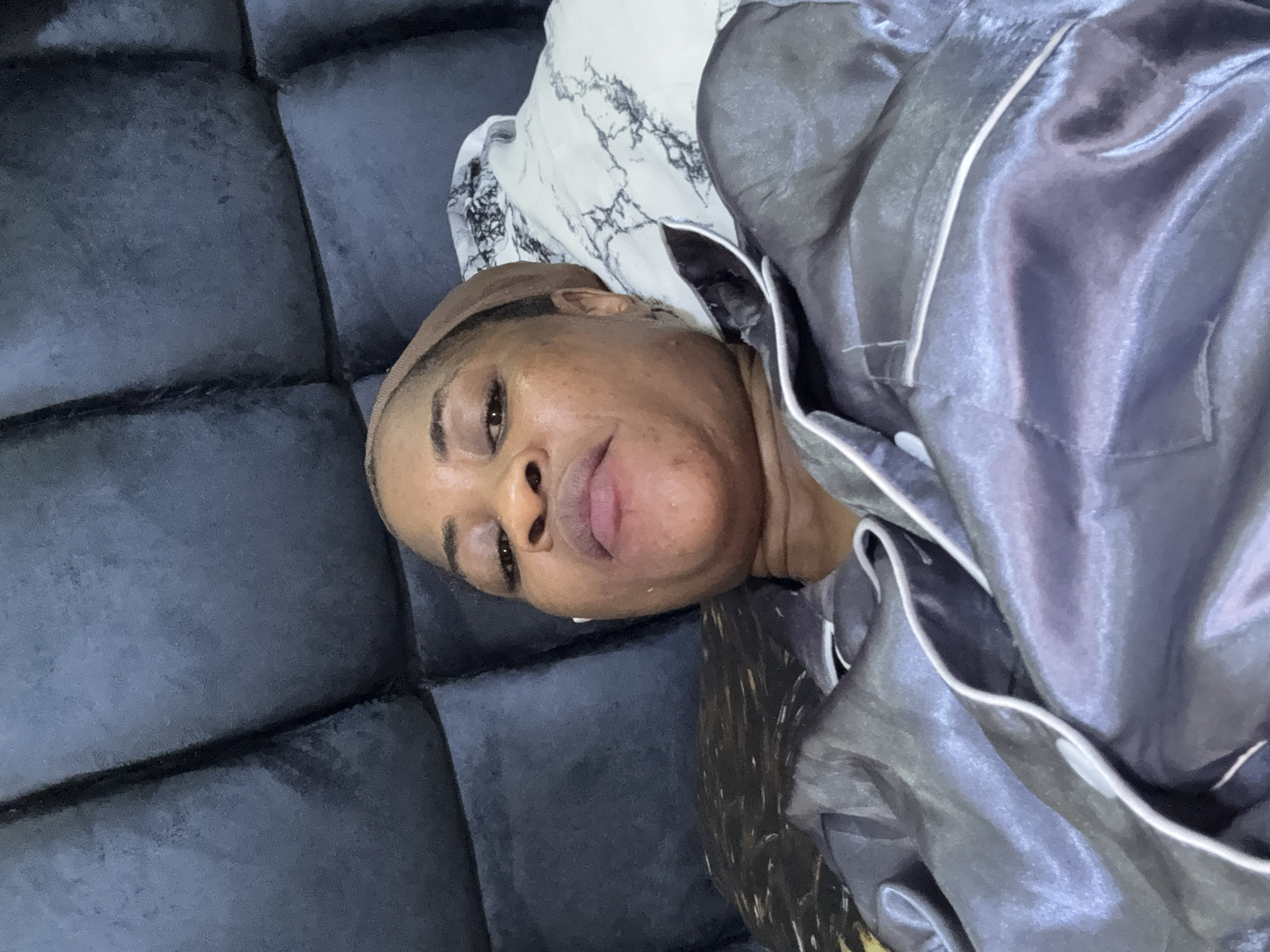 A dark-skinned woman with a brown head wrap is lying on her back in bed, wearing a grey satin pajama set with white piping. She is looking directly at the camera with a neutral expression. Her head rests on a pillow with a black and white marble pattern. The background consists of a dark, quilted headboard. The scene appears to be indoors, likely in a bedroom. The woman is positioned slightly off-center in the frame. There are no visible indications of the specific location within Ajara, Nigeria.