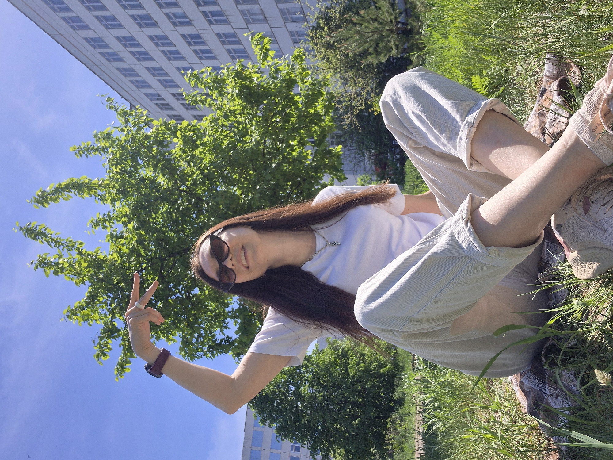A medium shot captures a woman relaxing in a grassy area within a city environment, her pose suggesting leisure and approachability. The woman, sporting sunglasses and a white t-shirt, is lying on the grass, with her legs slightly raised and bent. She is wearing light-colored pants rolled up at the cuffs, and her shoes are visible near her feet. She holds up her left hand in a "peace" sign, displaying a watch on her wrist. Behind her, lush green trees provide a natural backdrop against a clear blue sky. In the upper left and lower right corners of the image, segments of a tall, modern building with numerous windows suggest an urban setting. The location appears to be a park or green space within an urban environment, but the city is not specified.