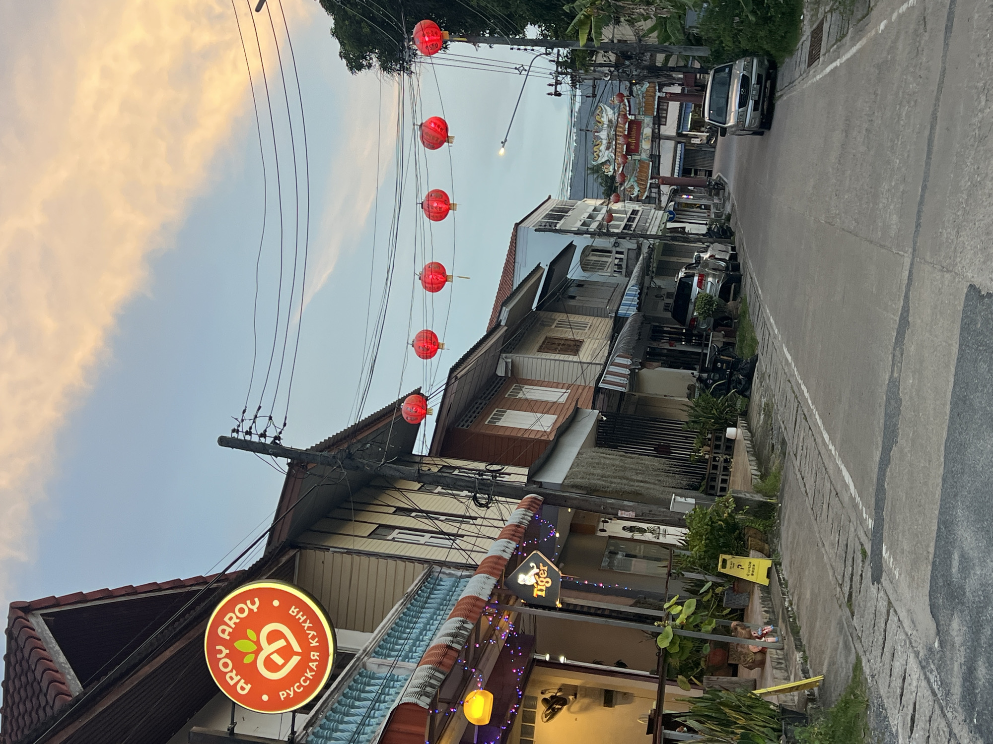 The image depicts a street scene in Baan Mae Nam, Thailand, at dusk. The sky is a gradient of light blue and soft orange hues, indicative of sunset, with scattered clouds. The street is paved with concrete, marked by a white line on the right side. Several buildings line the left side of the street, featuring traditional Thai architecture with wooden facades and tiled roofs. Some buildings have awnings and decorative lights. A prominent circular sign with an orange background, featuring the words "AROY AROY" and stylized heart and leaf graphics, is visible on one of the buildings. Below this, text in Cyrillic script reads "РУССКАЯ КУХНЯ," suggesting a Russian restaurant. Another sign with the "Tiger" logo is also visible. Red paper lanterns are strung across the street, suspended by wires, and are illuminated. A few vehicles, including a silver SUV and a white sedan, are parked along the roadside. Lush green plants in pots and planters are placed on the sidewalks and in front of some establishments. On the right side of the street, there are more trees and vegetation, and a distant ornamental archway, possibly a gateway to a Chinatown district, is visible. The overall atmosphere is one of a quaint, possibly tourist-oriented, street in a coastal town, transitioning from day to night.