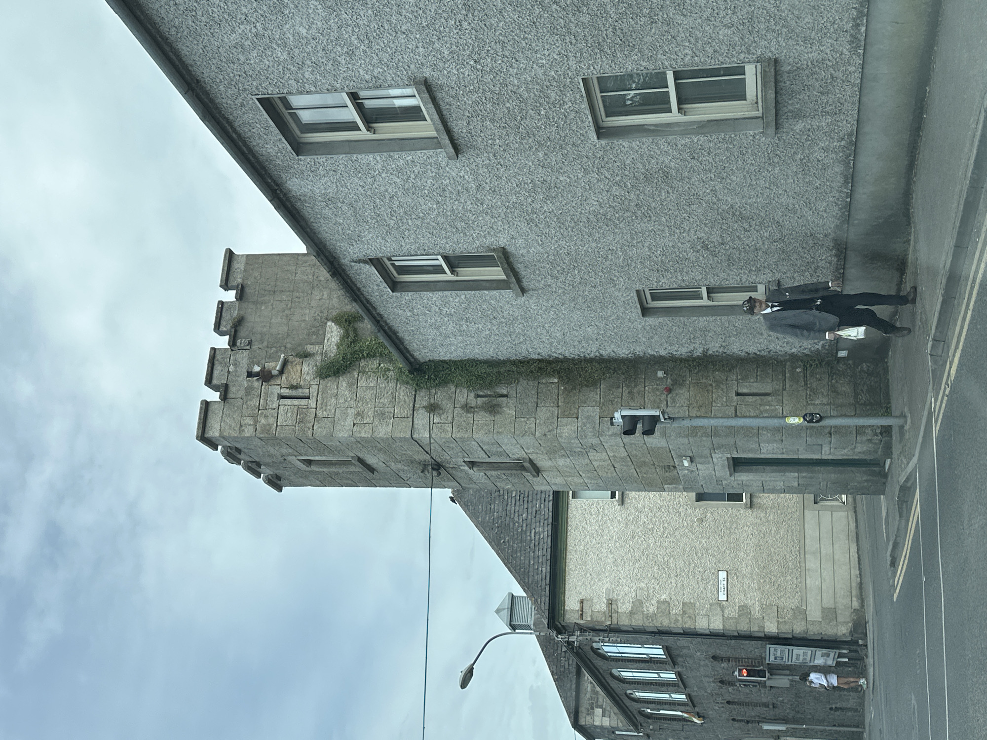 The image depicts a street scene in Carlow, Ireland, featuring a mix of architectural styles and a pedestrian. In the foreground, a man in a gray suit, dark tie, and a flat cap is walking on the sidewalk, carrying a white plastic bag. He is facing slightly towards the viewer. Behind him, a textured gray building with multiple windows lines the street. To the left of the building, a tall, stone tower with crenellations rises, possibly a historical structure, with ivy growing on its side. Further to the left, another building with arched windows is visible. A traffic light is positioned on the right side of the tower, currently showing red. In the background, the sky is overcast with scattered clouds. The street is paved with asphalt, marked with yellow lines. A street sign that reads "CHAFF ST" is affixed to one of the buildings. Another person, a woman in a light-colored dress, is standing near a traffic light and a signpost on the far left of the image.