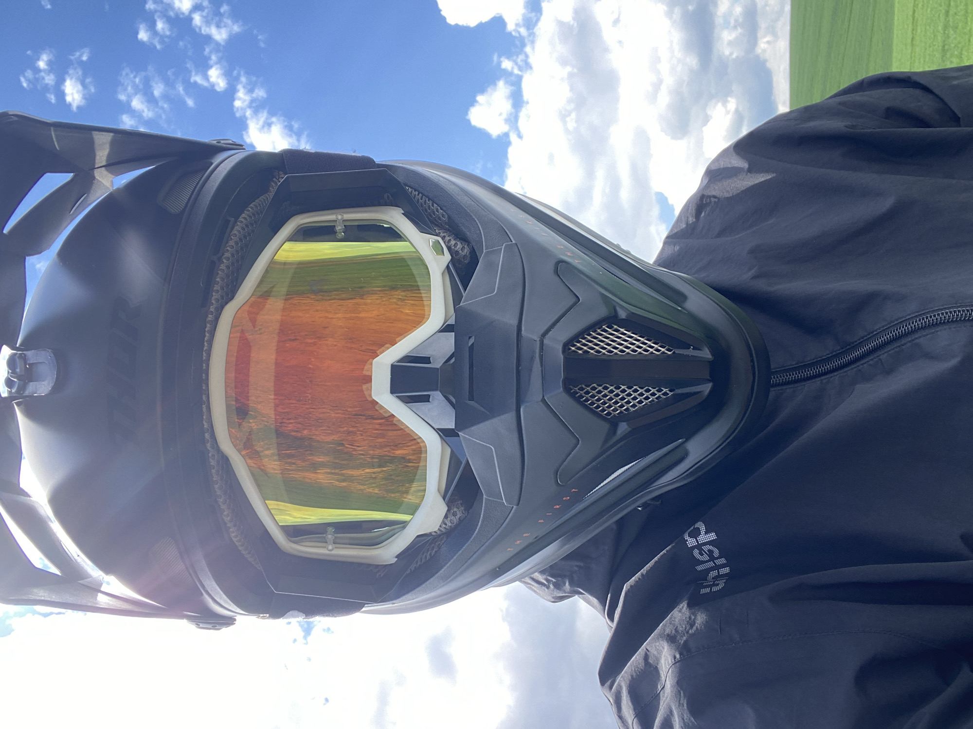The image presents a close-up view of a person wearing a black motocross helmet with orange-tinted goggles. The reflection in the goggles shows a field or grassy area under a partly cloudy sky. The person is wearing a black jacket with white lettering on the sleeve. The setting appears to be outdoors in a rural or semi-rural area. There is no definitive information in the image to establish it is in Beloslav, Bulgaria.