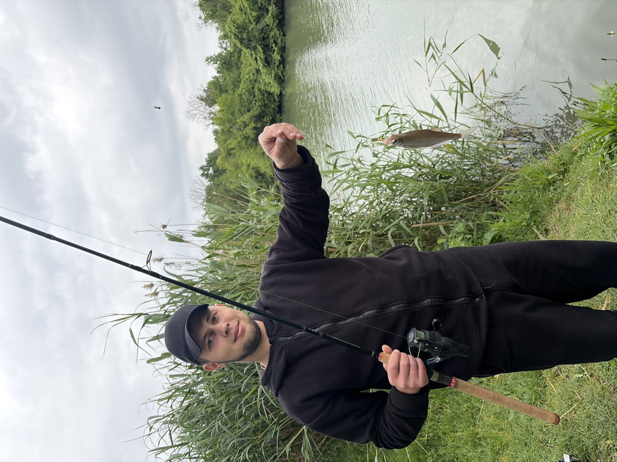 A man is seen in a full shot, holding a fishing rod and a fish in front of a body of water. The man is wearing a black jacket, pants, and a cap. The fishing rod is dark colored with a wooden handle and a reel. The fish is small, silver, and hanging from a line. The body of water is a river or lake, with green vegetation growing along the banks. In the background is a forest of green trees. The sky is overcast. Considering the greenery and the presence of a river or lake, this scene could potentially be within the vicinity of the Kuban River, which runs through Krasnodar, Russia. More specific location details are not discernible from the given image.