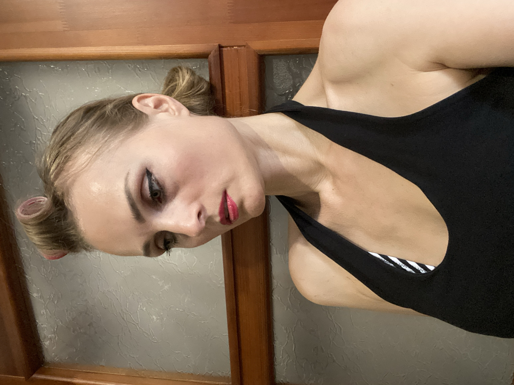 A Caucasian woman with blonde hair styled in pin curls and a bun, wearing a black halter-neck top with a striped bandeau underneath, poses for a selfie indoors. Her makeup includes winged eyeliner and bright pink lipstick. The background shows a textured wall and a wooden door frame, possibly in Lida, Belarus. The lighting is soft, casting subtle shadows.