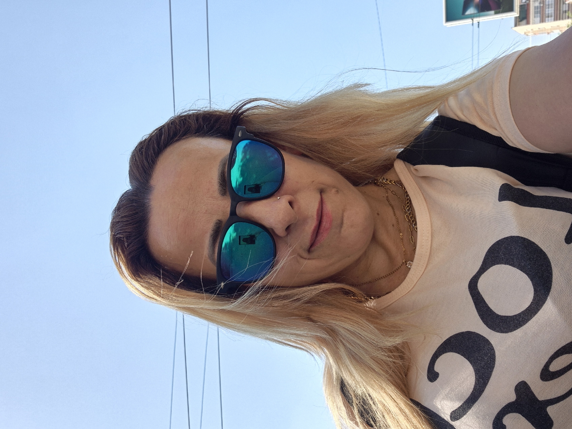 A low-angle, eye-level, close-up selfie of a light-skinned woman with long, blonde hair and blue-tinted sunglasses. She is wearing a light peach-colored t-shirt with large, dark gray lettering that reads "2000". She has a small nose ring and a mole on her right cheek. Two gold necklaces are visible around her neck. The background consists of a clear blue sky with a few thin, dark electrical wires running horizontally across the frame. To the right side of the image, a tall, illuminated billboard with a person on it is visible, along with the upper floors of a building. The lighting suggests it is daytime.