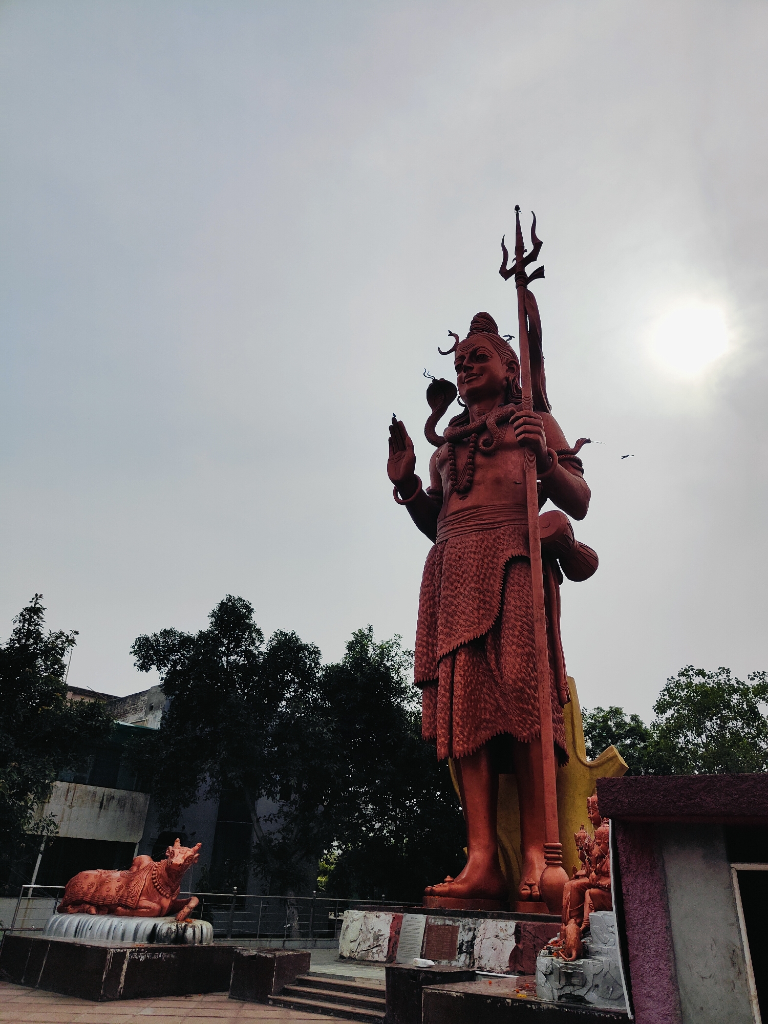 A low-angle, wide shot captures a towering, reddish-orange statue of Lord Shiva standing prominently against a hazy sky in Gurgaon, India. The statue is depicted in a standing pose, holding a trident in its right hand and a drum in its left. A snake is coiled around its neck, and a serpent also appears to be held in its left hand. The statue is adorned with a garland of beads and wears a patterned skirt. To the left of the main statue, a smaller, reddish-orange statue of a bull (Nandi) is seated on a pedestal. In the background, dark green trees and a light-colored building are visible. The sun is partially obscured by clouds, creating a diffused light. Several small birds are seen in the sky. The base of the main statue features other smaller statues and decorative elements, including a seated figure.