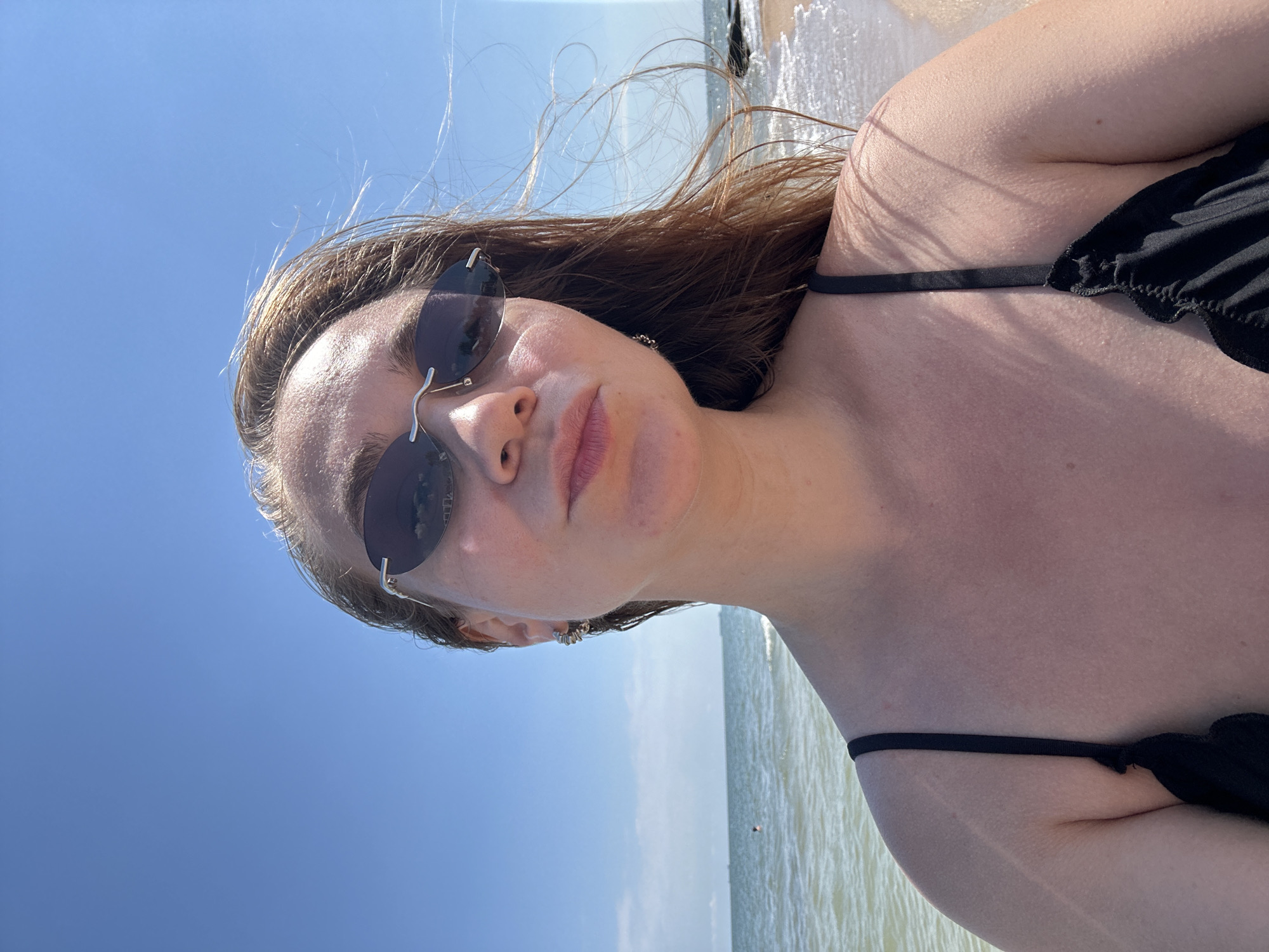 A young woman with long, brown hair, wearing sunglasses and a black bikini top, takes a selfie at the beach. Her hair is windswept. The sea and a blue sky are visible in the background. The location is Golubitskaya, Russia.
