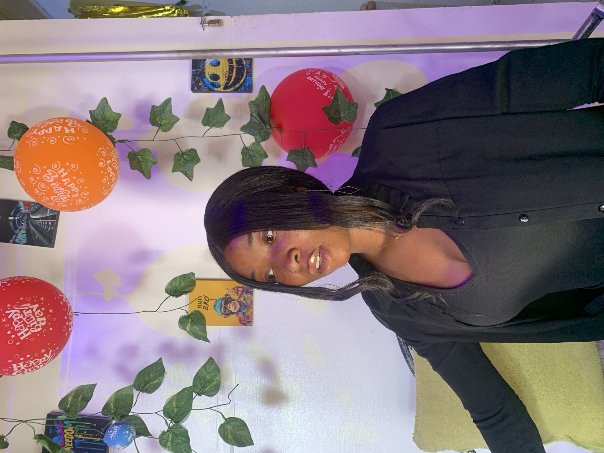 A woman is the main subject in this indoor shot, likely a self-portrait. She has dark hair and is wearing a black top and jacket. Her face is well-lit, allowing her features to be seen clearly.
The background consists of a white wall adorned with decorations, including several balloons printed with "Happy Birthday" messages. One balloon is orange, and two are red. Artificial green vines are draped across the wall, adding to the celebratory atmosphere. There are also posters or images tacked to the wall, though their exact content is difficult to determine due to the angle and image quality.
The scene appears to be a birthday celebration, possibly set up in a home or apartment. There is no specific information to confirm the location as Jos, Nigeria.