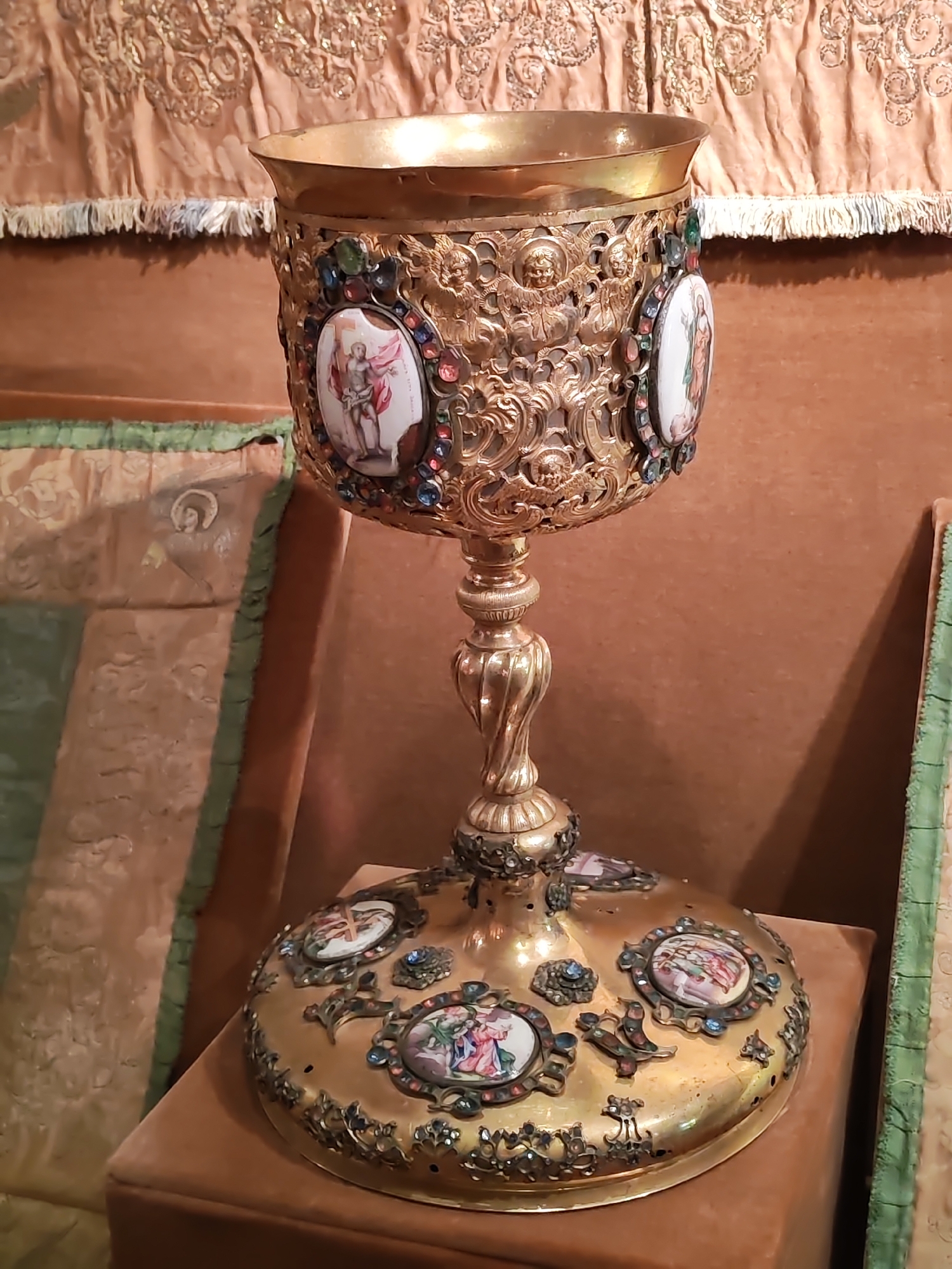 This is a close-up, eye-level shot of an ornate, golden chalice, likely made of brass or gilded metal. The chalice is elaborately decorated with numerous small, colorful gemstones and intricate metalwork depicting cherubs and scrollwork. Several oval medallions featuring painted religious scenes are inset into the cup and the base. On the cup, the medallions depict figures that appear to be saints. On the base, there are medallions with scenes that include people and possibly animals. The chalice stands on a twisted stem and a wide, circular base, also adorned with decorative elements. The chalice is placed on a cushioned surface, possibly a velvet pillow, which is a light brown or tan color. In the background, there is a richly textured, beige fabric with ornate embroidery and a fringed trim, suggesting a decorative tapestry or curtain. To the left and right of the chalice, glimpses of other upholstered objects with green trim are visible, hinting at a larger religious or museum setting. The lighting is soft and even, suggesting indoor lighting, and there are no indications of the time of day or weather. No visible text is present in the image. The overall impression is one of wealth, artistry, and religious significance, typical of liturgical objects or historical artifacts.