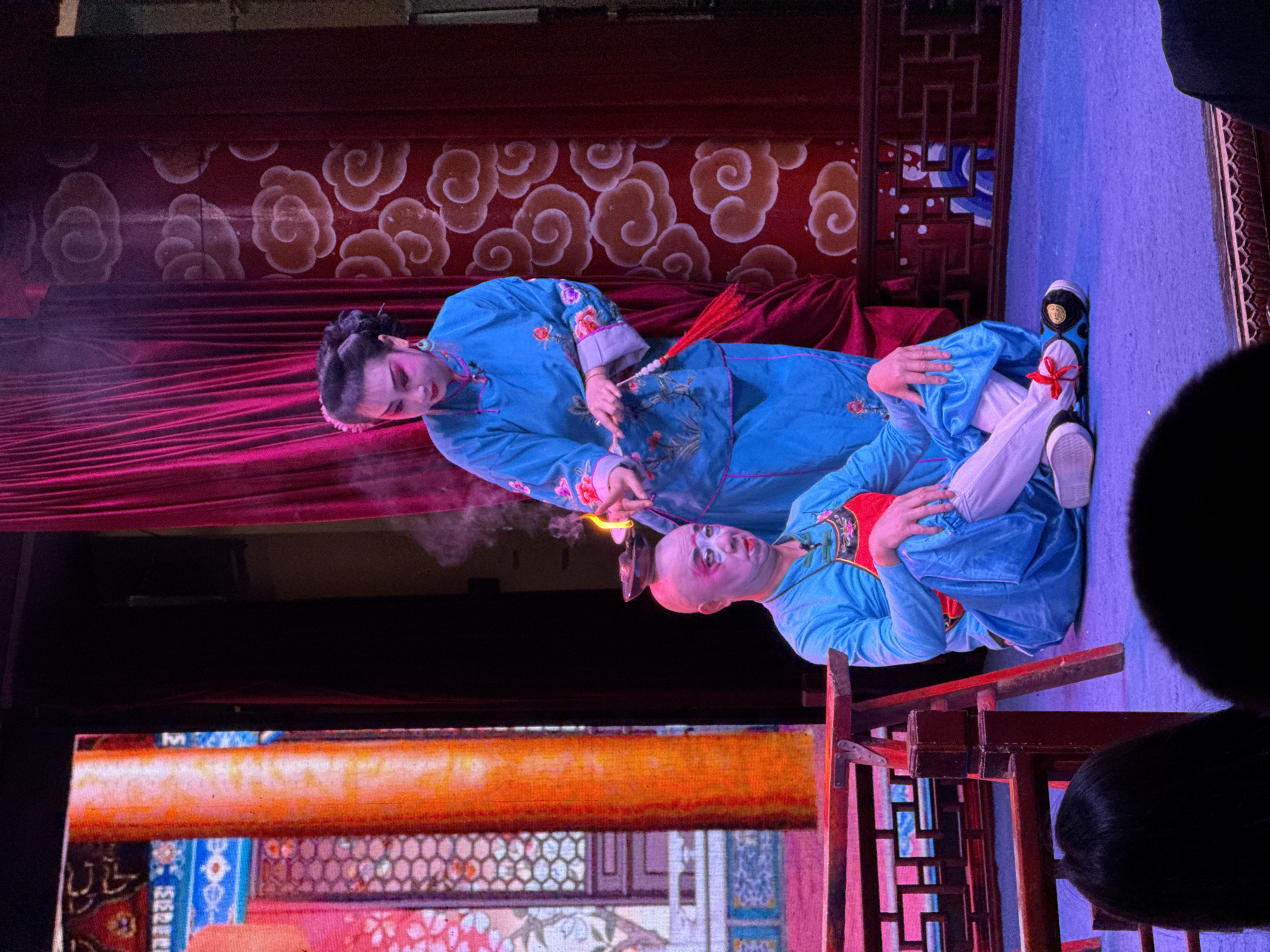 Two individuals in traditional Chinese opera attire are performing on a stage, likely in the Shapingba District, China. The performer on the left, dressed in a blue robe with floral embroidery and wearing traditional makeup, is holding a fan. The performer on the right, also in a blue robe, is lying on a blue surface. A wooden structure with intricate designs is visible at the bottom of the frame, and a screen displays vibrant patterns. The background features a red curtain with cloud-like patterns. A faint cloud of smoke or steam rises near the center of the image.