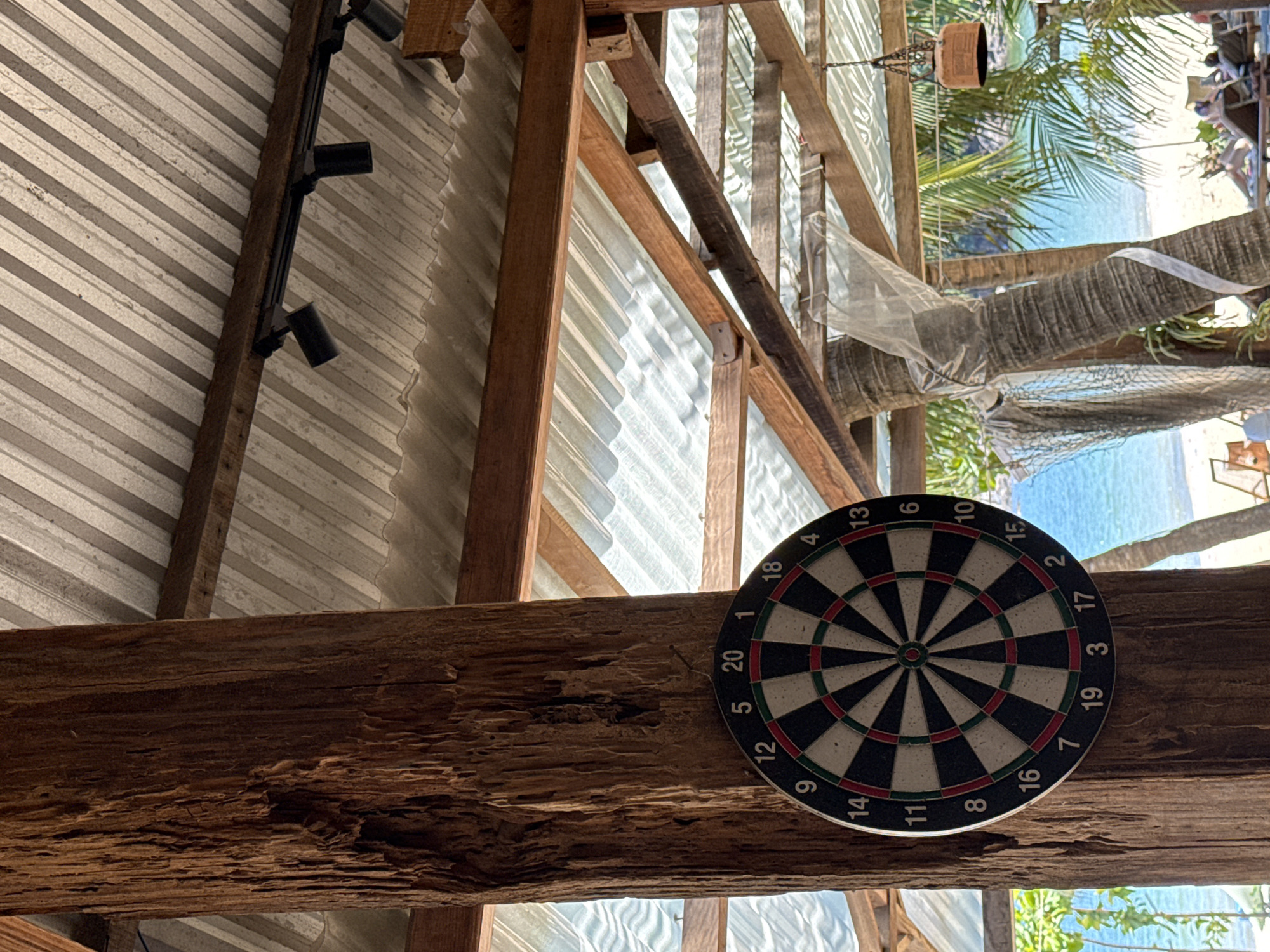 The image shows an indoor/outdoor space with a view of the beach, possibly in Đà Nẵng, Vietnam. A dartboard hangs on a wooden beam, which is part of the building's structure. The building has a metal roof and wooden framework. Outside, palm trees and the ocean are visible. Some decorative mesh is visible as well. There is also a spotlight in the background attached to the metal roof.