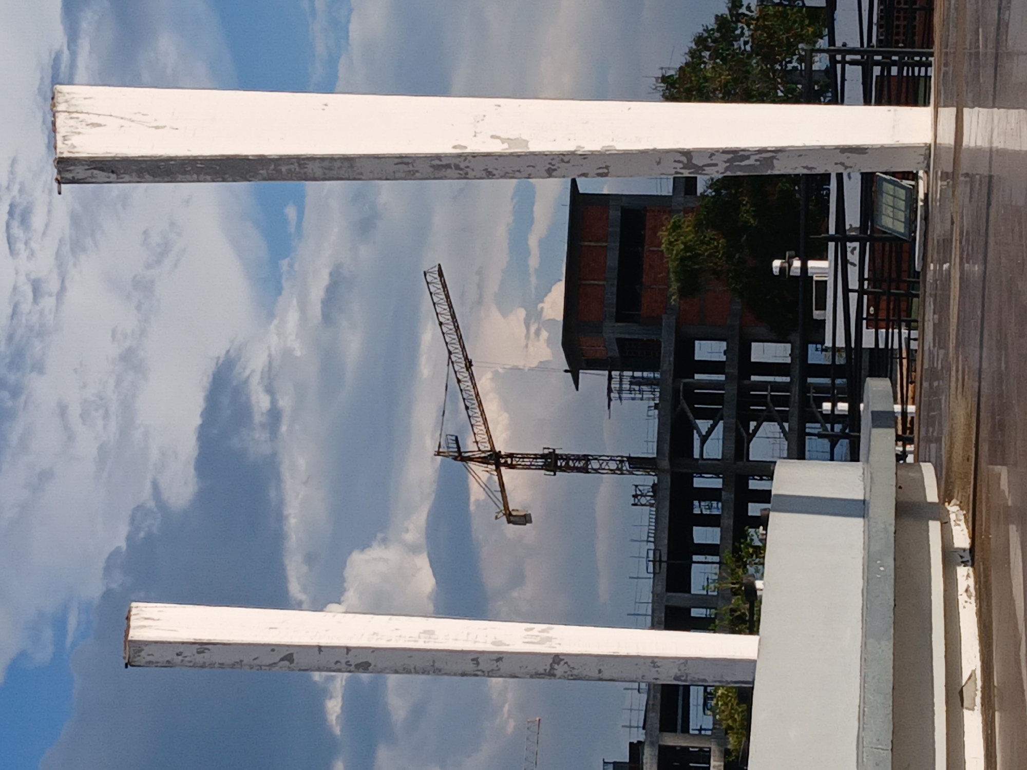 The image, which appears to be rotated 90 degrees clockwise, captures an outdoor scene in Ciudad Guayana, Venezuela, focused on a construction site. The foreground is framed by two weathered white horizontal beams, possibly concrete or painted metal, showing signs of peeling paint and discoloration. To the right, a textured grey concrete structure, perhaps a wall or pillar, is also visible.
The midground is dominated by a clear blue sky interspersed with fluffy white and grey clouds. Against this backdrop, a large yellow and black tower crane stands prominently, indicating active construction. Below and to the right of the crane, a multi-story building is under construction, featuring reddish-brown brickwork on some levels and exposed concrete or steel framework on others. Parts of the building appear to have dark, possibly reflective, facades. Green trees and bushes are scattered around the construction area. The scene depicts daytime under a bright, cloudy sky. No people or legible text are visible in the image.