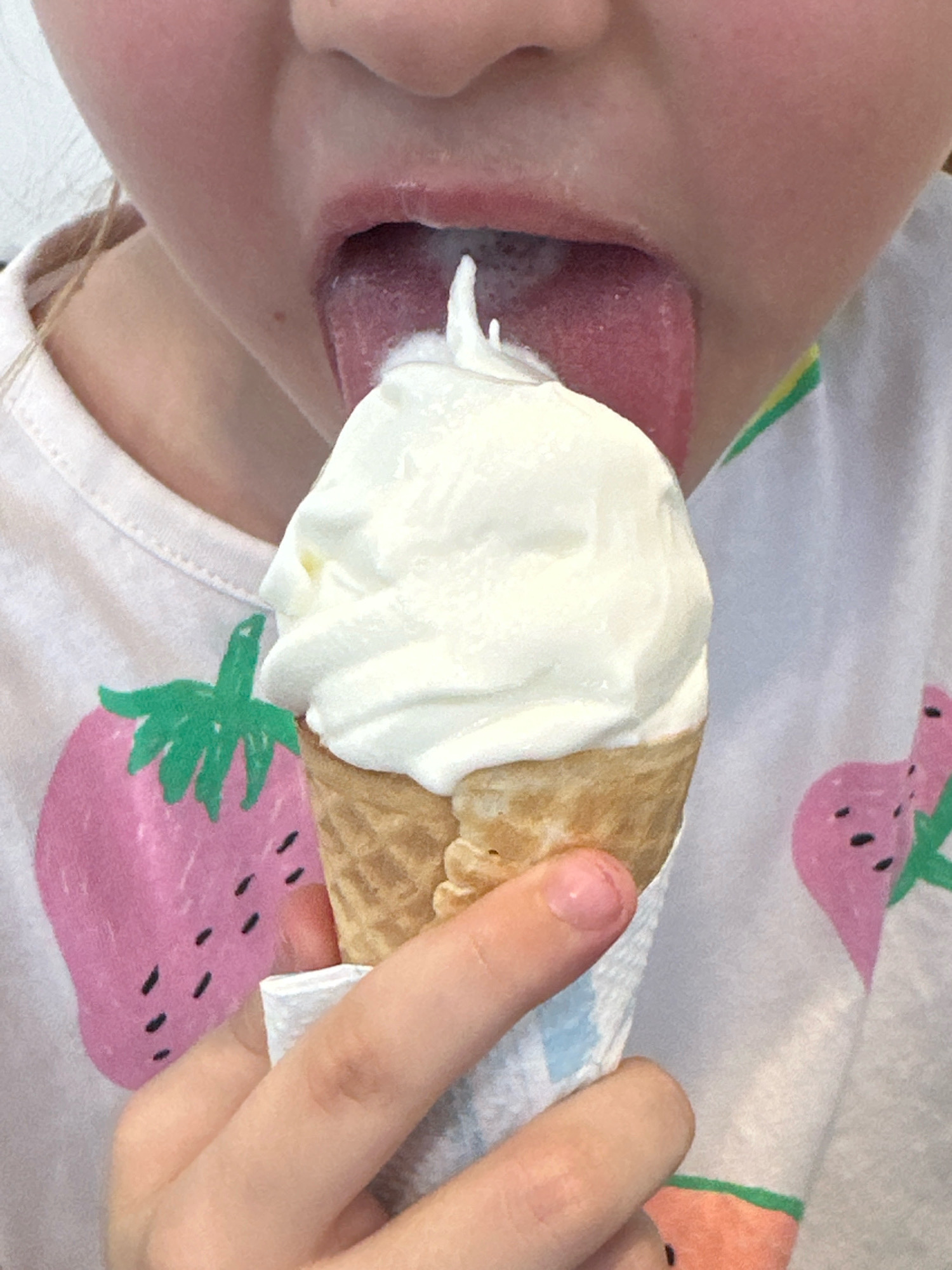 A close-up shot captures a person's face and hand as they enjoy an ice cream cone. The individual's mouth is open, with their tongue lapping at the soft-serve ice cream. The ice cream is white and swirled, perched atop a waffle cone. The person is wearing a light-colored t-shirt adorned with a colorful strawberry print. The background is out of focus, providing no contextual information about the location in Rio de Janeiro, Brazil. The lighting suggests it is daytime.