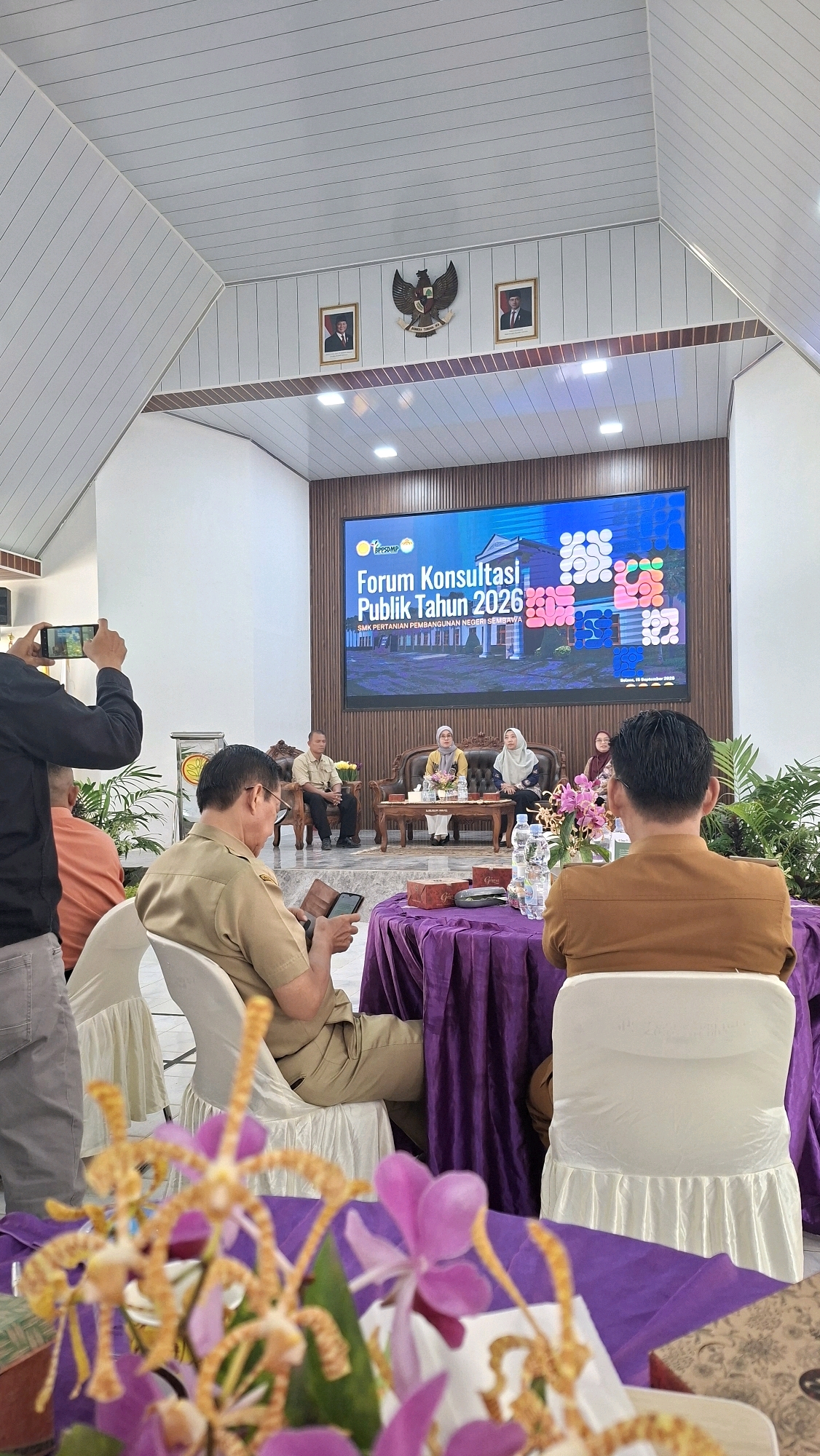 In a bright, modern indoor venue in Lalang Sembawa, Indonesia, a forum consultation is underway, as indicated by the large LED screen displaying "Forum Konsultasi Publik Tahun 2026" and "SMK Pertanian Pembangunan Negeri Sembawa." Four individuals are seated on a low, ornate sofa on a stage area, facing the audience. A man in a light brown uniform, likely a government official, is seated to the left, looking down at his phone and holding a small book or planner. To his right, a woman in a yellow hijab and a grey cardigan is speaking, gesturing with her hand. Next to her, another woman in a darker hijab is seated, listening. A third woman in a patterned hijab is also seated on the sofa. In front of the stage, an audience is seated, facing the speakers. A man in a tan uniform is in the foreground, seated and looking at his phone. To his left, another person in an orange shirt is visible, and further to the left, a person is taking a photo of the stage with their phone. The room has a white, sloped ceiling with recessed lighting. Two framed portraits of men and an eagle emblem are mounted on the wall above the stage. Tables draped in purple fabric are set up for the audience, with water bottles and floral arrangements. White chairs with fabric skirts are arranged for the attendees.