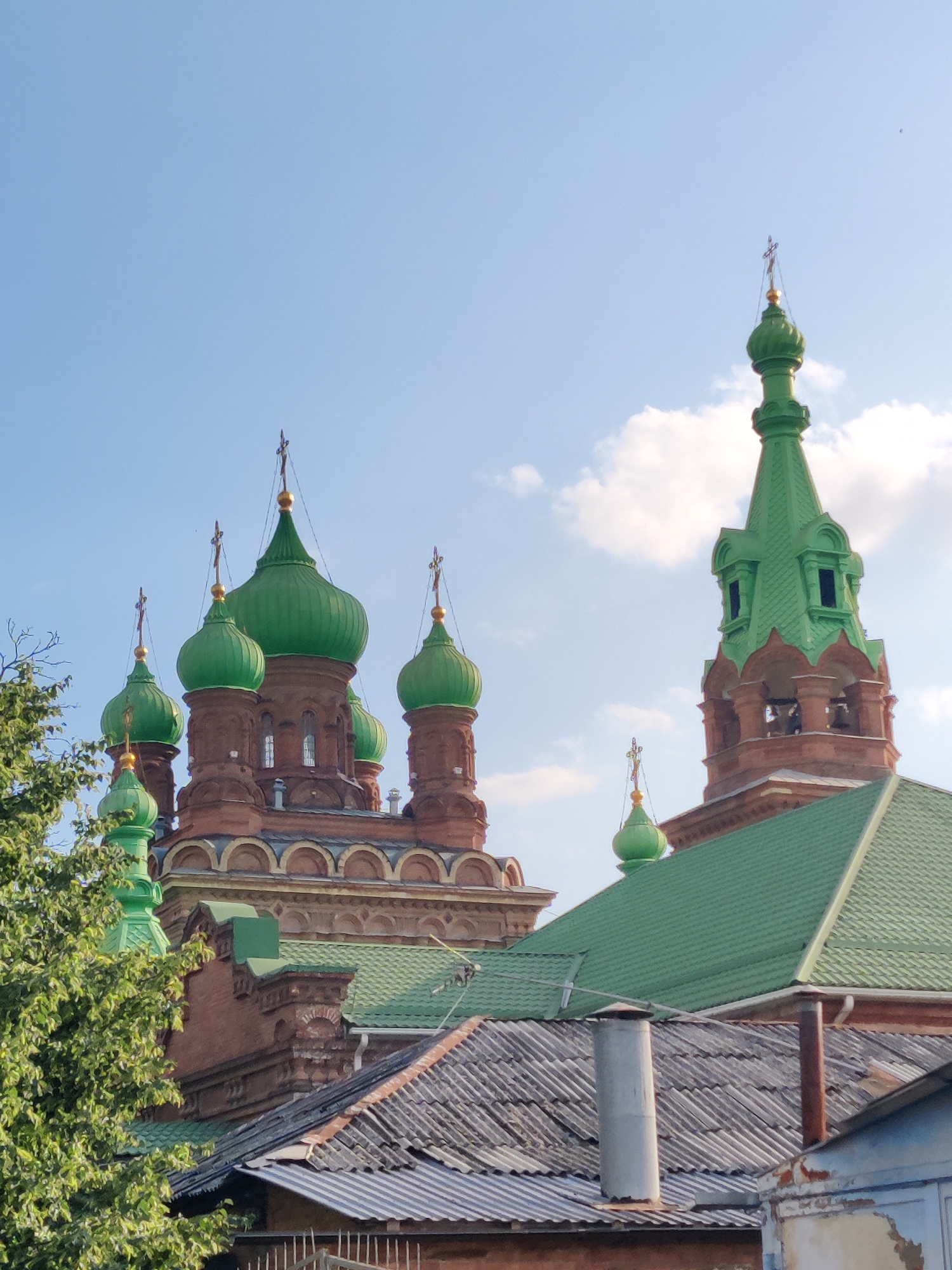 The image displays a view of the Saint Nicholas Orthodox Cathedral in Krasnodar, Russia. The cathedral is characterized by its numerous green domes and reddish-brown brickwork. A tall, slender bell tower with a green roof and open arched sections is visible on the right side of the image, also adorned with green elements and a golden cross at its apex. Several smaller green domes, topped with golden crosses, punctuate the roofline of the main building. In the foreground, the rooftops of less ornate, older buildings with corrugated metal roofs are visible, partially obscuring the lower portions of the cathedral. A lush green tree with dense foliage is situated on the left side of the frame, reaching towards the cathedral's domes. The sky above is a clear blue with scattered white clouds. The scene captures a sunny day, with the architecture of the religious building being the primary focus.