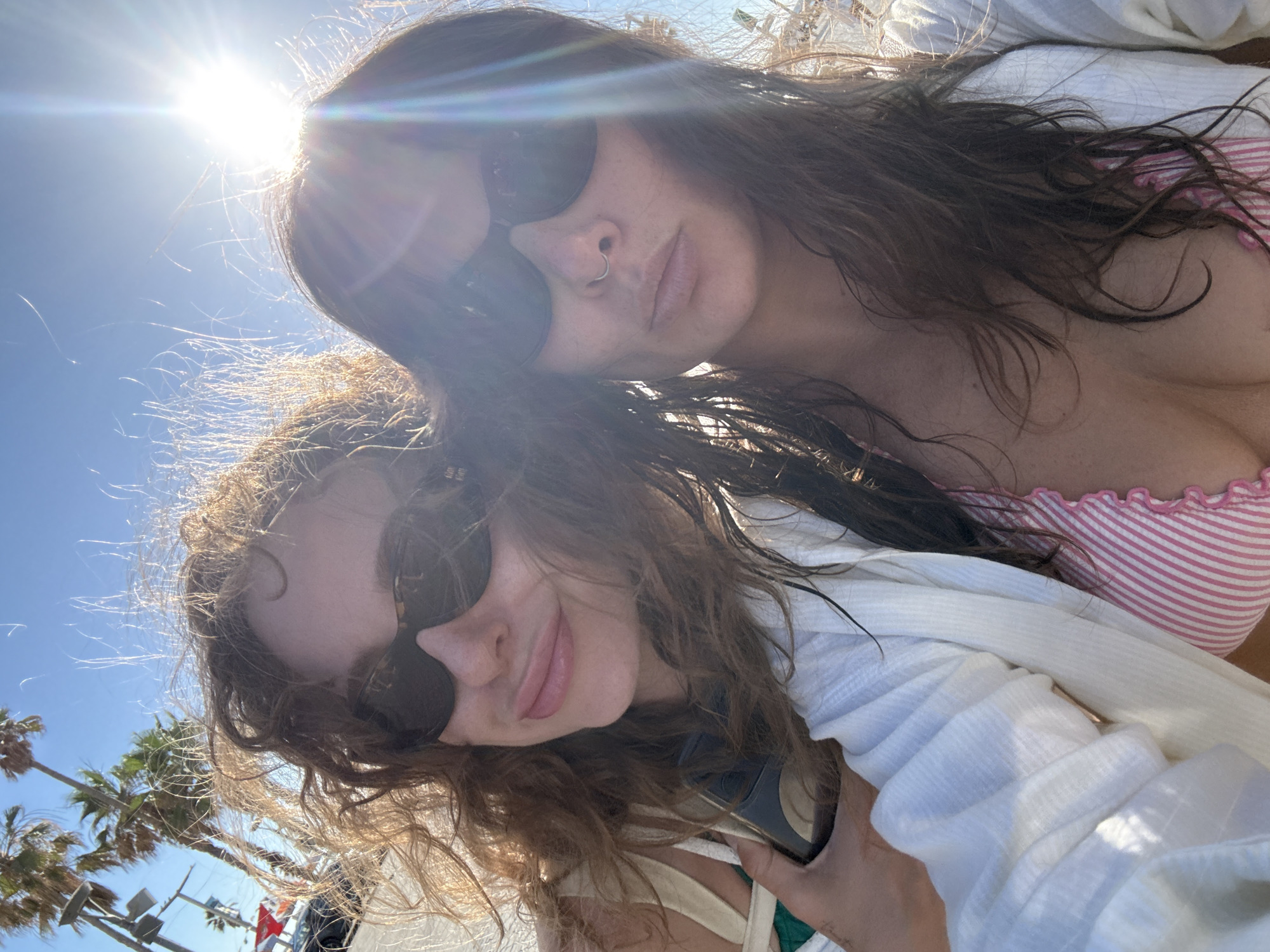 Two women are taking a selfie outdoors. The woman on the left has long, curly brown hair and is wearing sunglasses. The woman on the right has long, dark brown hair and is also wearing sunglasses and a nose ring. She is wearing a pink and white striped bikini top and a white cover-up. The background shows palm trees, a bright sun with lens flare, and a clear blue sky. The image appears to have been taken on a sunny day. There is no clear indication of the location being Tel Aviv, Israel.