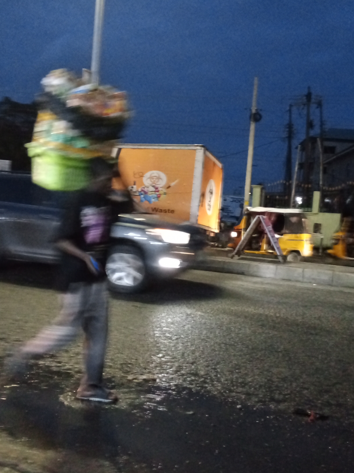 A man is walking on a wet street in Ado, Nigeria, carrying a large basket of items on his head. The basket contains various objects, including what appears to be green produce and other goods. He is wearing a black t-shirt with a purple design and grey pants. In the background, there is a large orange truck with "Waste" written on the side, suggesting a waste management context. To the right of the truck, a yellow tricycle is parked, with a ladder propped against it. The scene appears to be taking place at dusk or early evening, with streetlights and vehicle headlights illuminating the wet road. The overall environment suggests an urban setting with traffic and commercial activity.