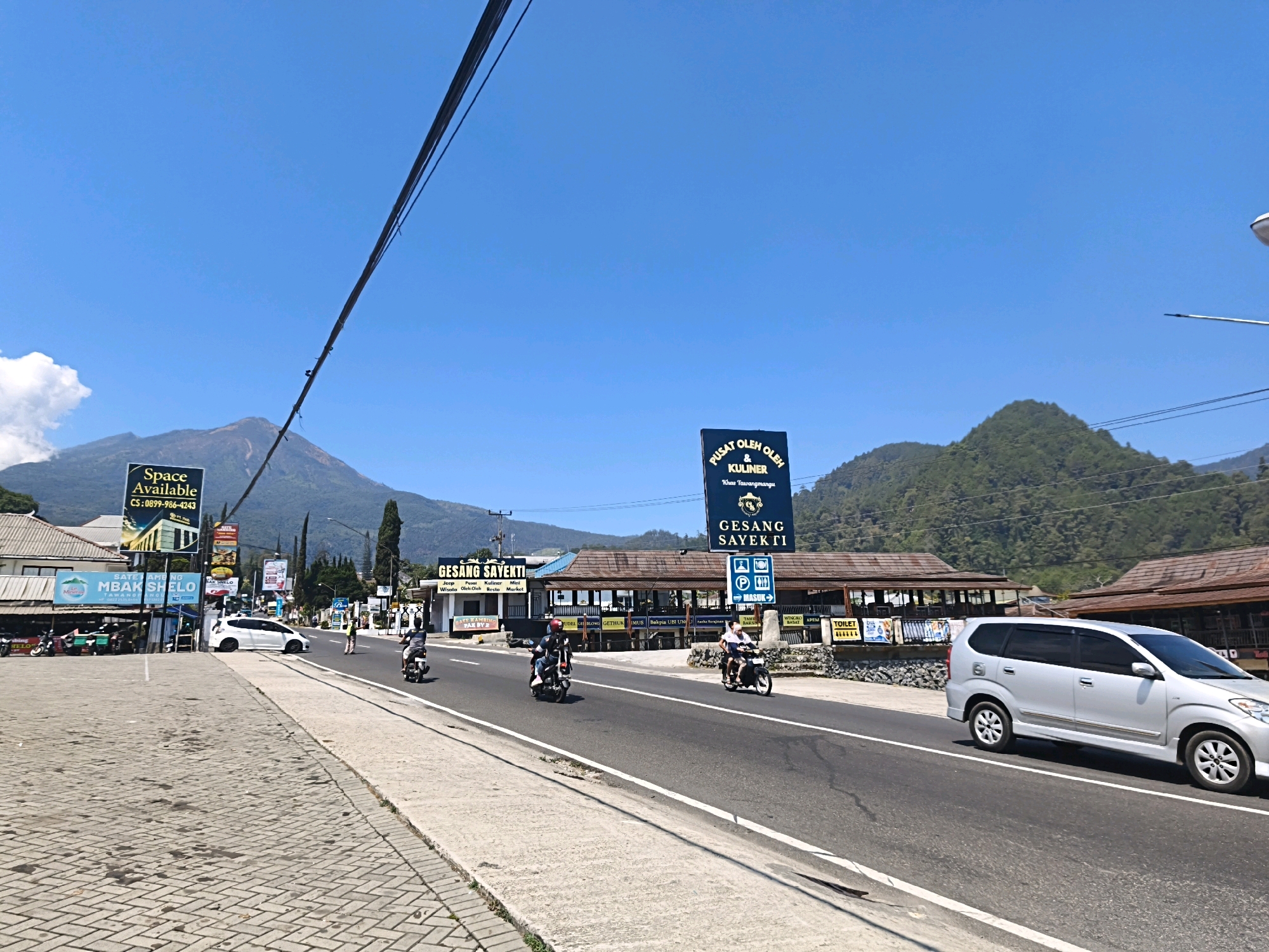 The image depicts a sunny day in Tawangmangu, Indonesia, with a prominent view of Mount Lawu in the background. The foreground shows a paved sidewalk bordering a two-lane asphalt road. Several motorcycles and a silver minivan are traveling on the road. On the left side of the road, there are buildings with signage, including one that says "Space Available." On the right side, there are more buildings with awnings, and signs for "GESANG SAYEKTI KULINER" (Gesang Sayekti Culinary) and "PUSAT OLEH OLEH & KULINER" (Souvenir & Culinary Center). These signs indicate that the area is a hub for local food and souvenirs. The overall scene is typical of a busy roadside in a tourist area.