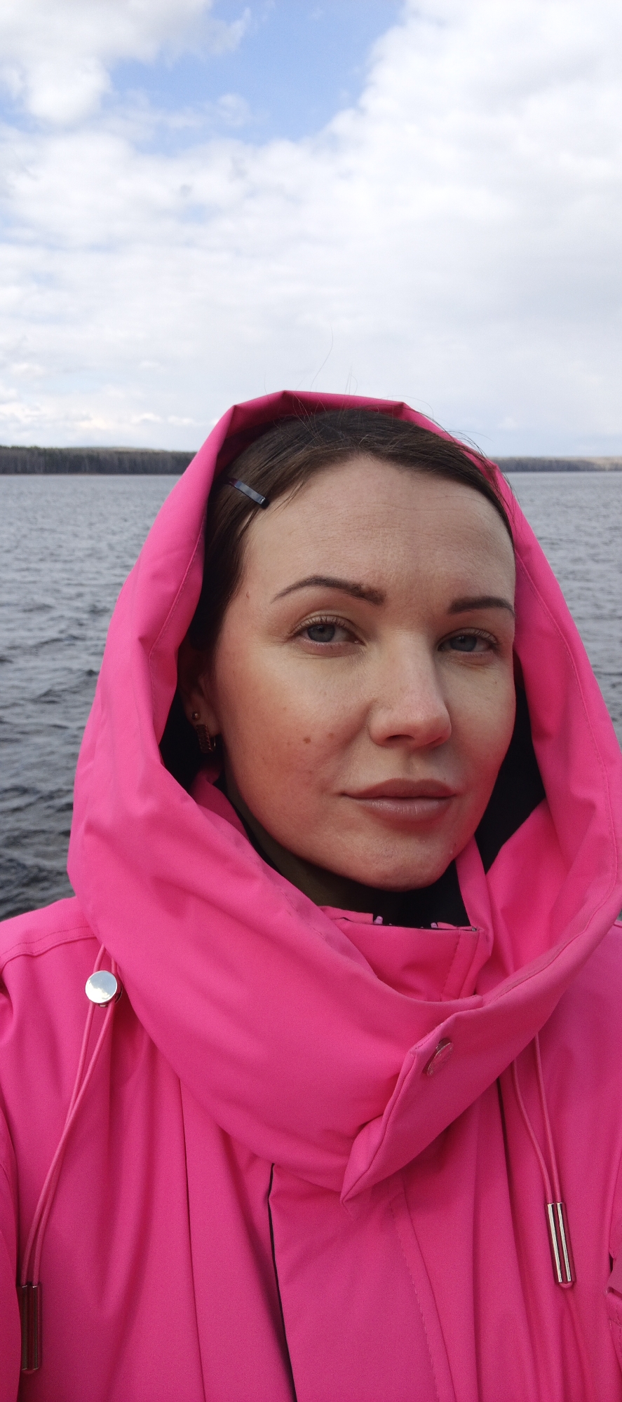 A woman poses for a selfie in front of a body of water on an overcast day. She wears a bright pink hooded jacket. Behind her is a dark body of water with a distant treeline. The sky is cloudy. The location is likely in or near the city of Chaikovsky, Russia, based on the prompt.