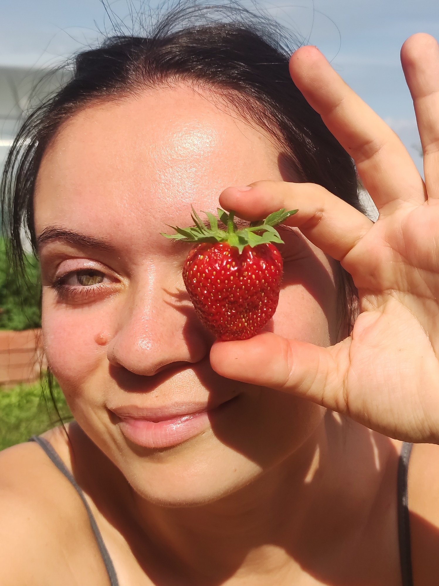 A close-up, selfie-style portrait shows a young woman holding a single strawberry in front of her right eye. Her left eye is visible and appears to be green or hazel. Her skin is illuminated by sunlight, highlighting her features and creating shadows. She has dark hair, some of which is falling forward. Her lips are slightly parted in a gentle smile. She is wearing a gray tank top. The background is slightly blurred, showing glimpses of greenery and what appears to be a structure, possibly a greenhouse, against a clear blue sky. The overall impression is one of a casual outdoor activity, likely in a rural or suburban setting, given the natural elements. There is no visible indication that this image was taken in the city of Фоки, Russia; the scene suggests a general outdoor environment.