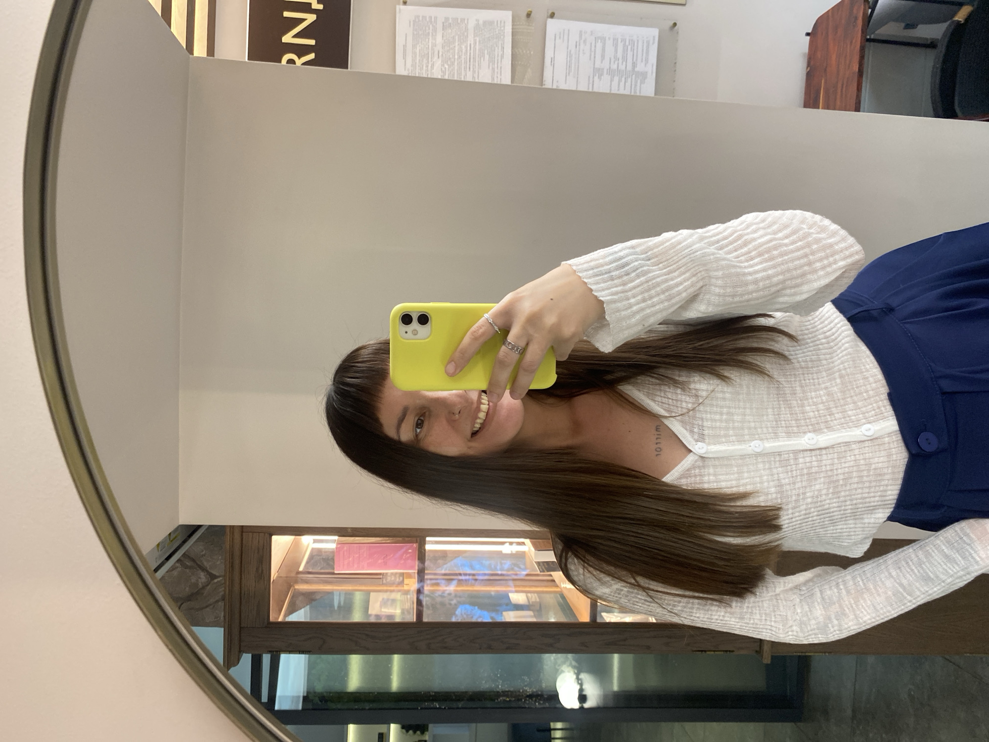 A woman with long, dark hair and bangs takes a mirror selfie indoors. She is smiling and holding a bright yellow smartphone in her right hand. She is wearing a white, ribbed, long-sleeved top with buttons down the front and a dark blue pleated skirt. A small tattoo is visible on her chest. The background includes a wooden display cabinet filled with items, a white wall, and a dark wooden table with a chair behind it, suggesting an indoor commercial or retail space. The scene is consistent with an establishment in Votkinsk, Russia, given the context.