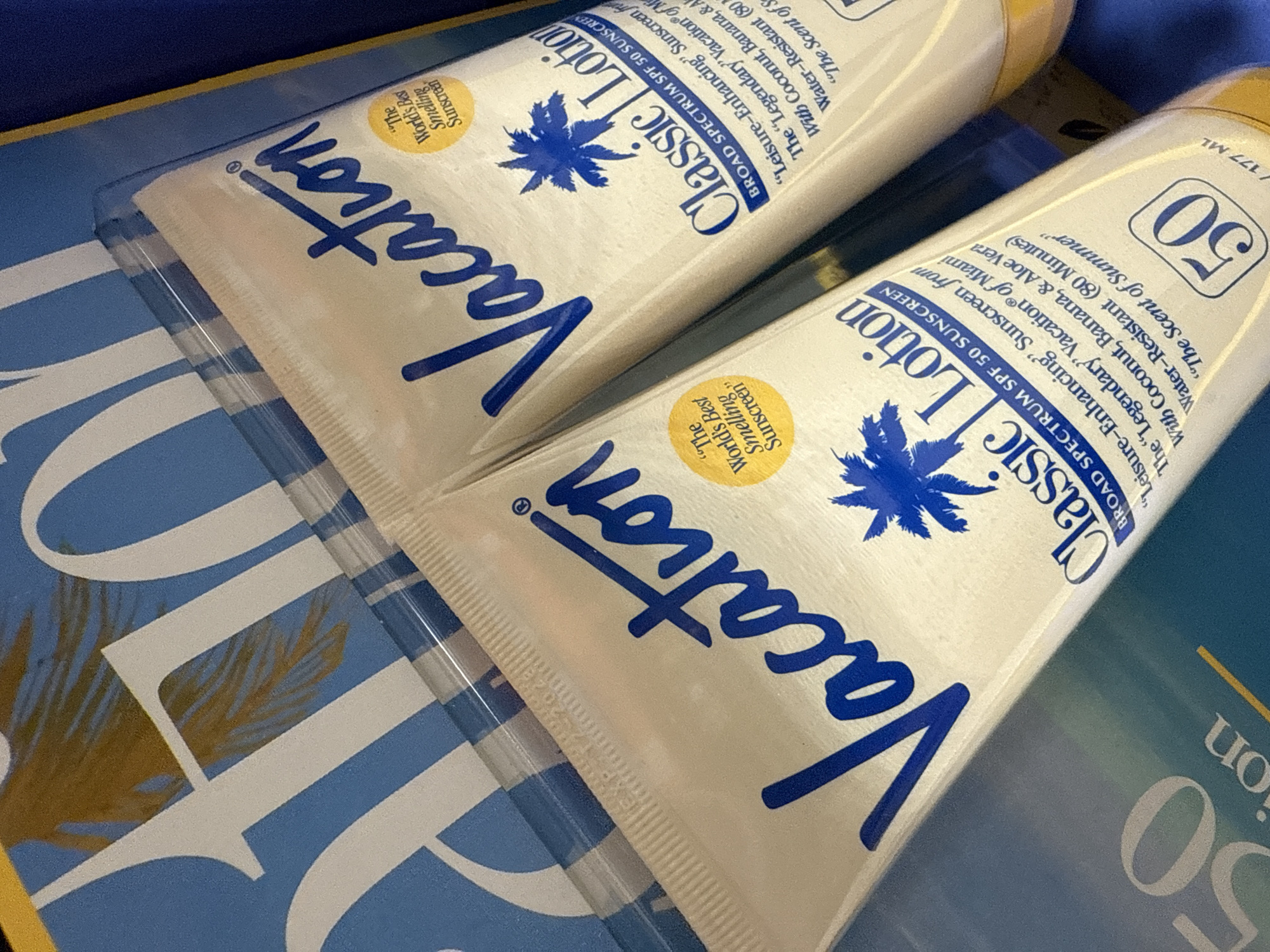 The image shows two tubes of "Vacation Classic Lotion" sunscreen with SPF 50. The tubes are white with blue lettering and a blue palm tree logo. The text on the tubes includes phrases like "Broad Spectrum SPF 50 Sunscreen", ""Leisure-Enhancing" Sunscreen from The "Legendary Vacation® of Miami"", ""The Scent of Summer"", "Water-Resistant (80 Minutes)", "With Coconut, Banana, & Aloe Vera", and ""The World's Best Smelling Sunscreen"". The tubes are placed on a blue and white patterned surface. The scene appears to be indoors and provides no indication of a location within Davie, United States of America.