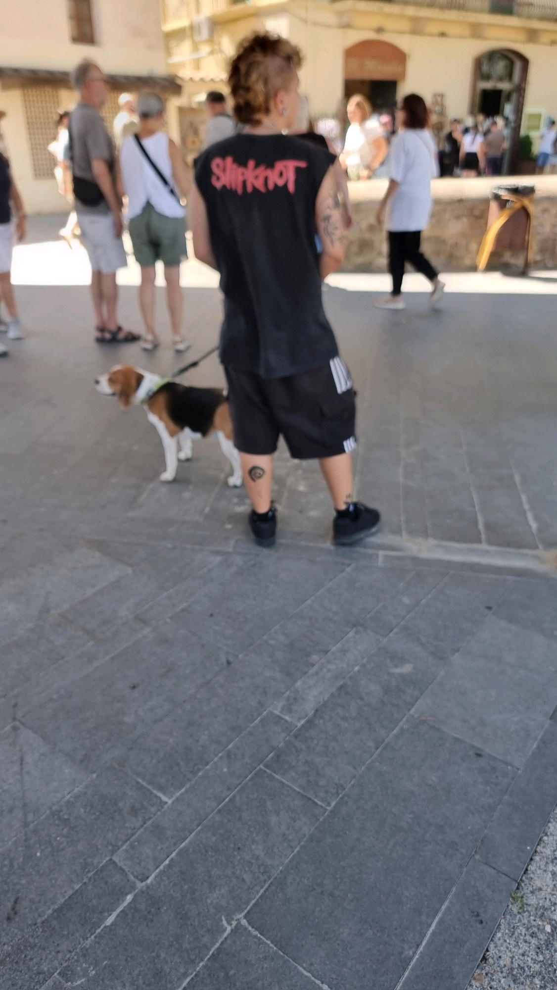 A person with a mohawk hairstyle and a black sleeveless t-shirt with the "Slipknot" logo in red is walking a beagle dog on a leash. The person is wearing black shorts with white stripes on the sides and black sneakers. A circular tattoo is visible on the person's right calf. The scene appears to be outdoors on a paved walkway, likely in the city of Besalú, Spain, given the architectural style of the buildings in the background. Other people are visible in the background, some walking, and a woman in a white shirt and black pants is walking in the middle ground. The environment is bright and sunny.