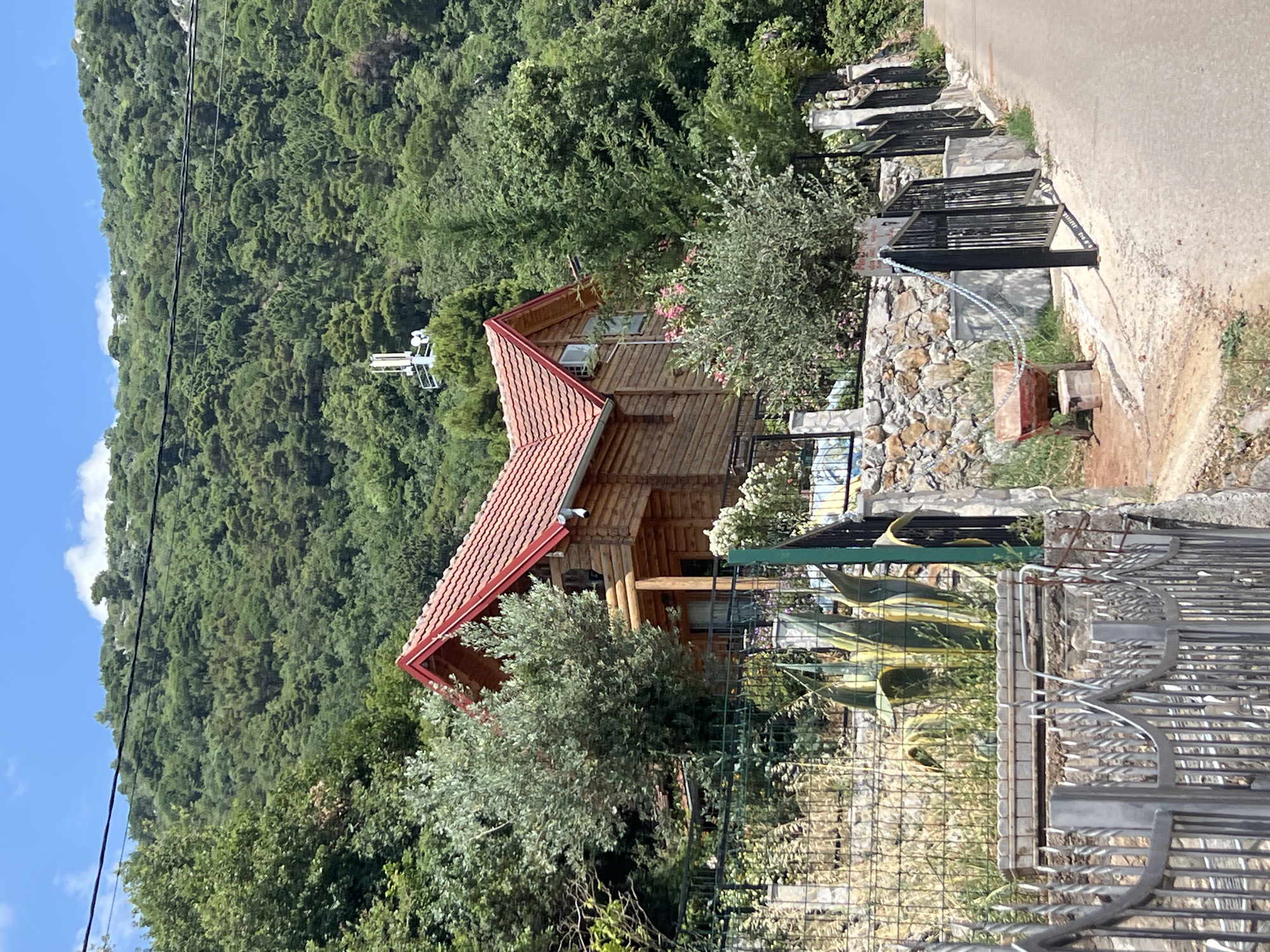 A rustic wooden cabin with a red tiled roof sits nestled on a densely forested mountainside under a clear blue sky. The cabin, constructed from horizontal logs, features several windows. In front of the cabin, a well-maintained garden with a fence and large agave plants is visible. A gravel road runs alongside the property, bordered by a stone wall and black metal fencing. In the distance, the verdant mountain slopes are covered in thick green trees. The sun appears to be high in the sky, indicating a daytime scene. There is no discernible activity or event taking place, and no text is visible in the image. The setting suggests a peaceful, natural environment, likely in a rural or mountainous area.