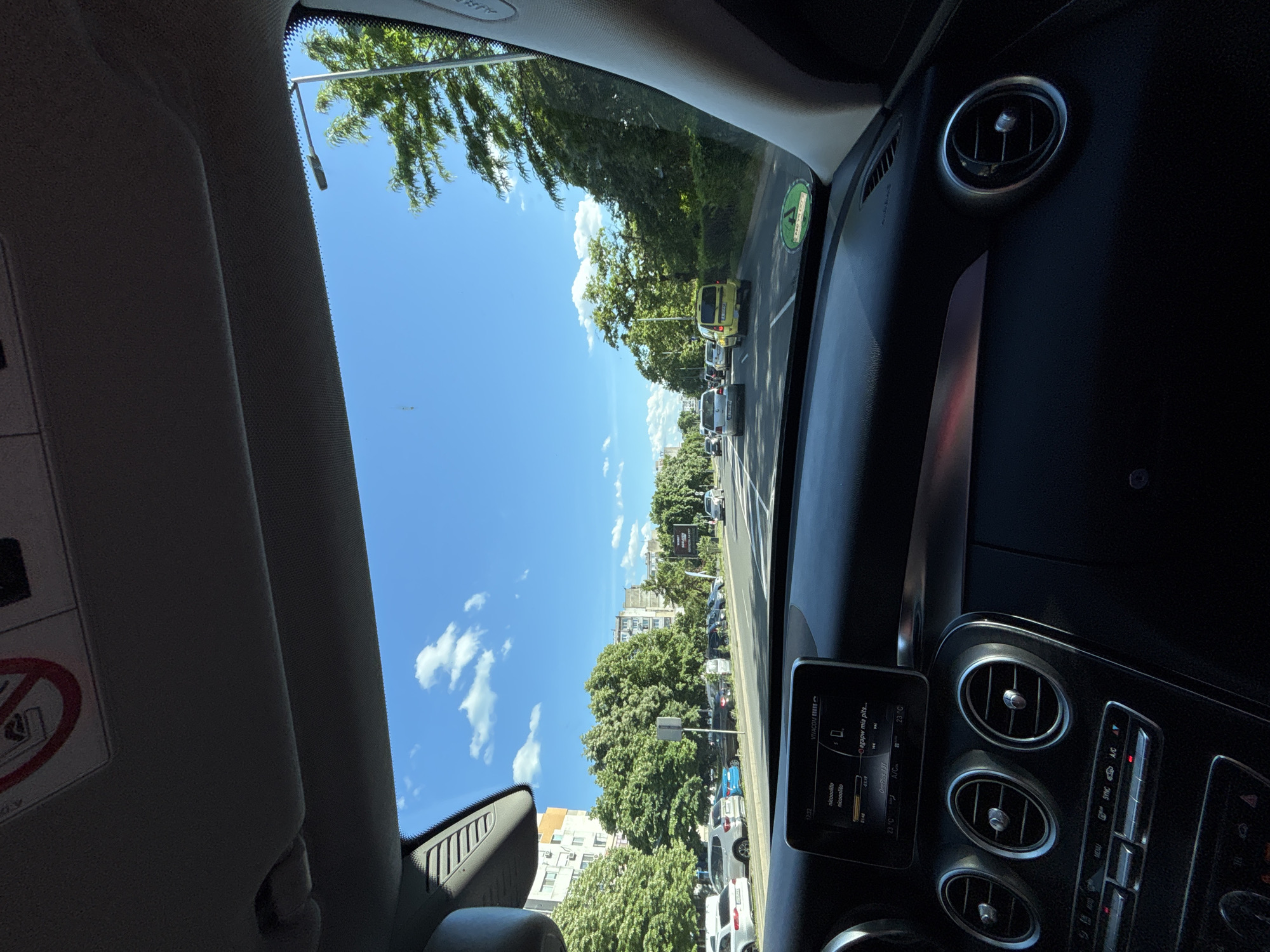 The image shows a view from inside a car, likely a Mercedes-Benz, looking out onto a street. The car is in a line of traffic on a sunny day. The sky is blue with scattered clouds. Trees line the street on both sides. Buildings are visible in the distance. A road sign and a yellow van are also visible in the distance. The car's dashboard is visible, with air vents and the car's multimedia display visible. Based on the information on the multimedia display, which includes the text "nicoolita nicocolito", it is possible that this image was taken in Burgas, Bulgaria.