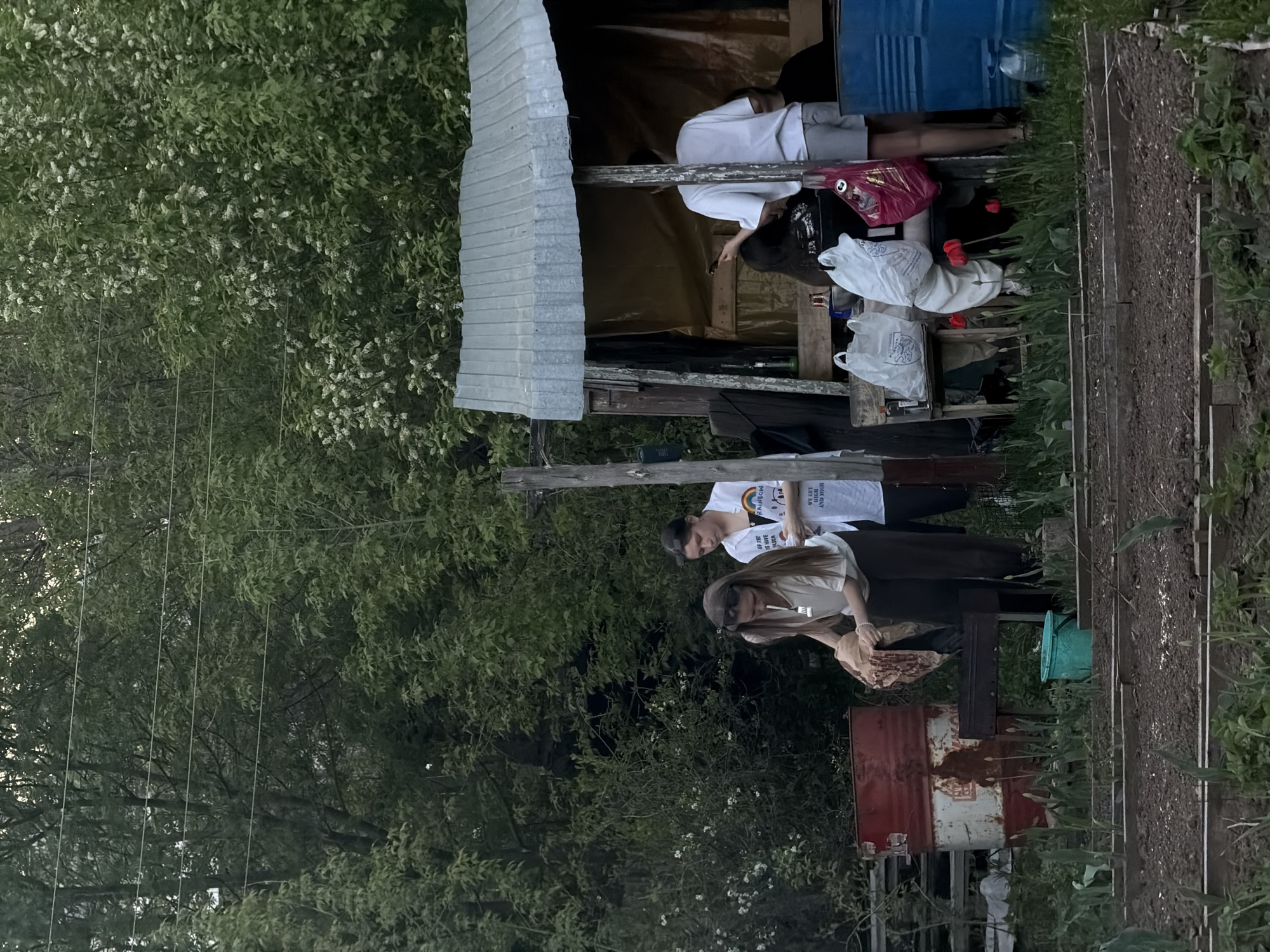 The image shows a scene in Perm, Russia, with two people in the foreground and another person resting in a makeshift shelter in the background.
Foreground:
Two people are standing near a garden area. One person, with long hair and wearing sunglasses, is holding something in their hands. The other person is wearing a white t-shirt with a rainbow graphic and words that appear to say "We get by with a little help". A metal barrel is also present, partially obscured by vegetation.
Background:
A makeshift shelter with a corrugated metal roof is visible. A person is lying inside the shelter. Bags and other items are also present inside and near the shelter.
Environment:
Trees and vegetation surround the scene. The overall environment appears to be a garden or outdoor area.