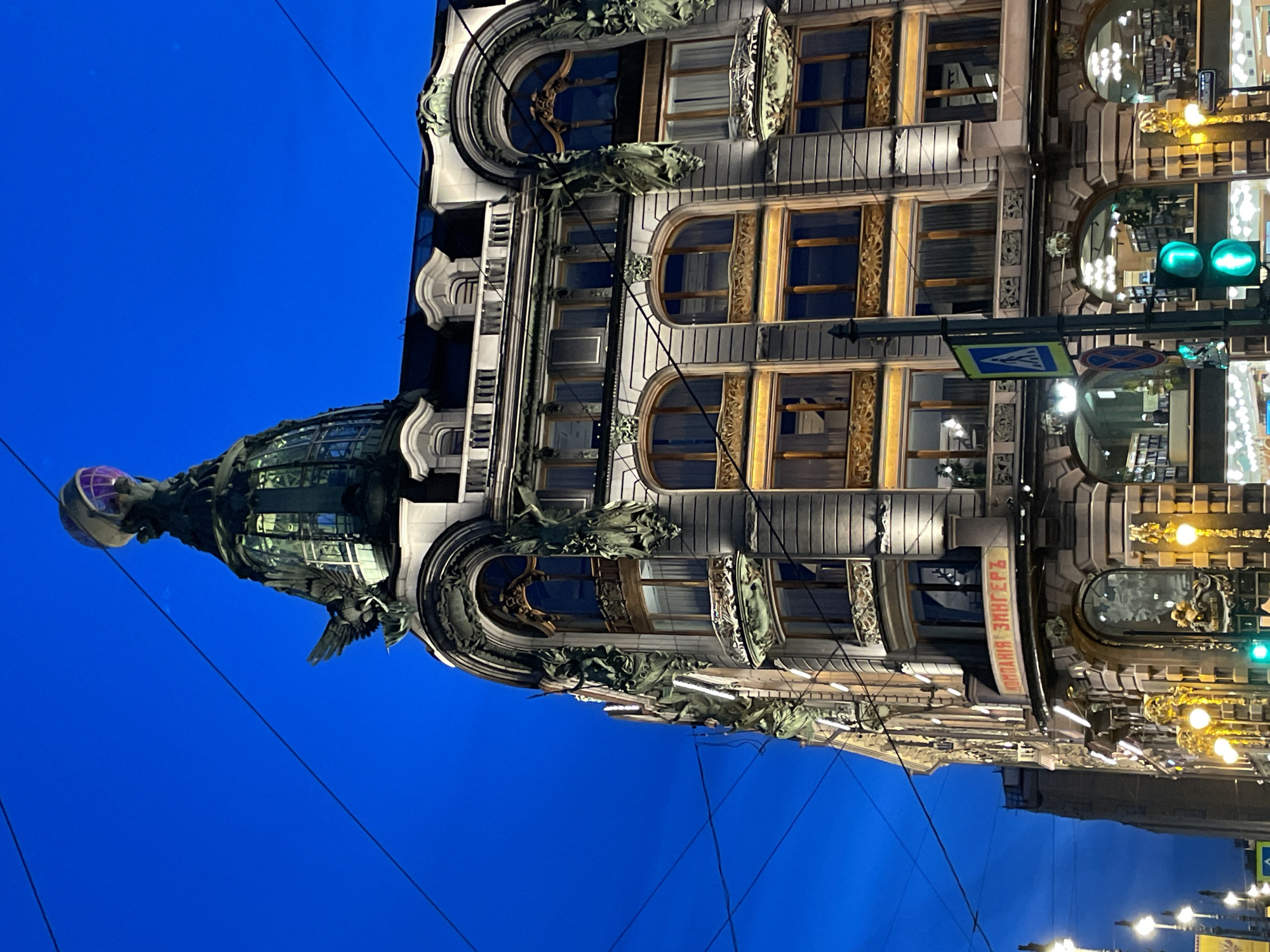 The image depicts the illuminated Singer House (Dom Knigi) in Saint Petersburg, Russia, at twilight or early night. The sky is a deep, clear blue, crisscrossed by overhead utility wires. The ornate, multi-story building, a prominent example of Art Nouveau architecture, is brightly lit by external floodlights, highlighting its intricate facade.
A large glass dome crowns the building, topped by a dark, sculpted eagle holding a spherical globe that emits a purple internal light. The facade features numerous decorative elements, including sculpted figures, arched windows, and balconies with intricate railings. A sign displaying "КОМПАНИЯ ЗИНГЕРЪ" (Company Singer) in illuminated red Cyrillic script is visible above the ground floor.
The ground floor's large arched windows reveal brightly lit interiors with bookshelves, indicating a bookstore. On the street level, a black pole supports a blue pedestrian crossing sign and a traffic light showing green for both pedestrians and vehicles. Warm yellow streetlights are visible on the left and right. No individuals are clearly visible on the street, though faint human figures can be seen inside the illuminated ground-floor establishments.