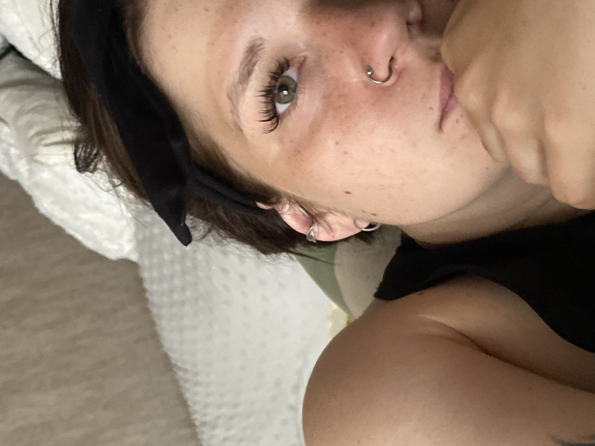 A close-up, slightly angled, eye-level shot shows a young woman lying on a bed. Her face is partially obscured by her hand resting on her chin. She has brown hair pulled back with a black headband, and her eyes are green with long, dark eyelashes. She is wearing a black top and has piercings in her nose and ear. The background consists of a textured white comforter and a beige wall, suggesting an indoor setting. The image does not contain any specific geographical markers to indicate its location within the city of Chaikovsky, Russia.