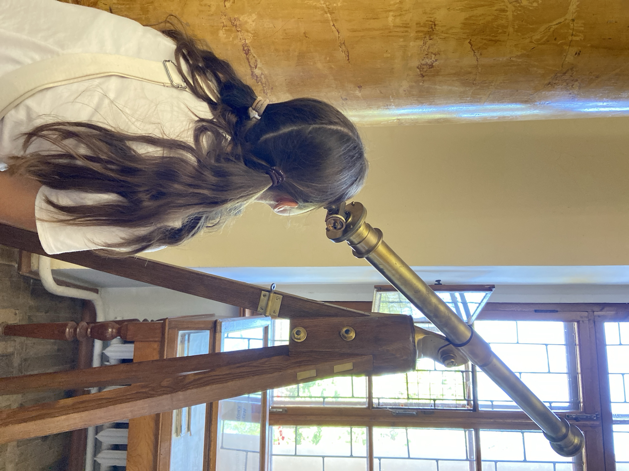 A young girl with dark brown hair in two pigtails is looking through a brass telescope mounted on a wooden tripod. She is wearing a white t-shirt. The telescope is pointed towards a large window with stained glass panes. To the left of the girl is a large, textured, golden-colored column. In the background, behind the telescope, is a wooden display case with glass doors, which sits on a wooden table with carved legs. The floor appears to be tiled or made of stone. The scene is indoors.