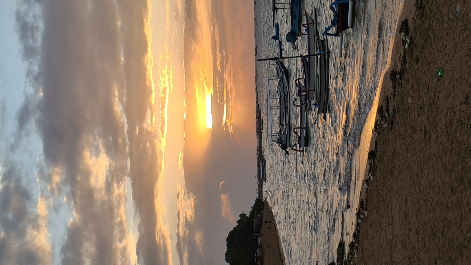 The image depicts a beach scene at sunset in Denpasar, Indonesia. The sky is filled with dramatic clouds tinged with orange and yellow from the setting sun, which peeks through a break in the clouds, casting a golden light onto the water. Several traditional Balinese fishing boats, known as jukungs, are anchored in the calm sea, their silhouettes visible against the illuminated horizon. The foreground is a sandy beach, with some scattered debris, indicating it may not be a heavily maintained tourist area. In the distance, along the coastline, a line of trees marks the edge of the land. A few people can be seen walking on the beach. The overall atmosphere is serene and picturesque, characteristic of a tropical sunset.