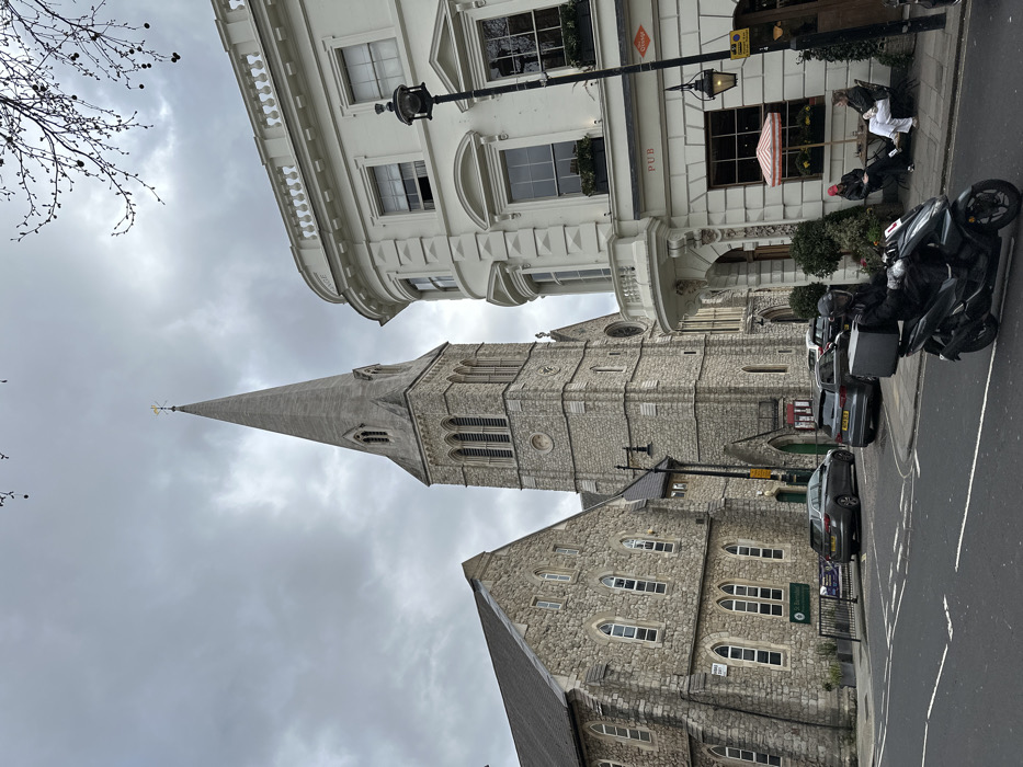 The image depicts an urban street scene in Pimlico, London, United Kingdom, under an overcast sky. On the left, St. Barnabas Church, a large stone-built structure, features a prominent grey spire capped with a golden weather vane and a lower stone building connected to its side, exhibiting numerous arched windows. To the right, "The Orange" pub, a three-story, light-colored building, displays classical architectural elements, including a curved corner with a small balcony and a "PUB" sign on its facade.
In the foreground, a black delivery scooter, ridden by a helmeted individual wearing dark clothing and carrying a large black delivery box on the rear, travels from right to left on the asphalt road. White road markings are visible on the street surface. Along the curb adjacent to the church, several cars are parked, including a dark grey station wagon and another dark grey sedan. Outside The Orange pub, two individuals are seated at an outdoor table beneath a red and white striped umbrella. One person wears a red hat, while the other is dressed in light-colored attire. Bare tree branches extend into the upper right portion of the frame. Street lamps and various signs are present throughout the scene.