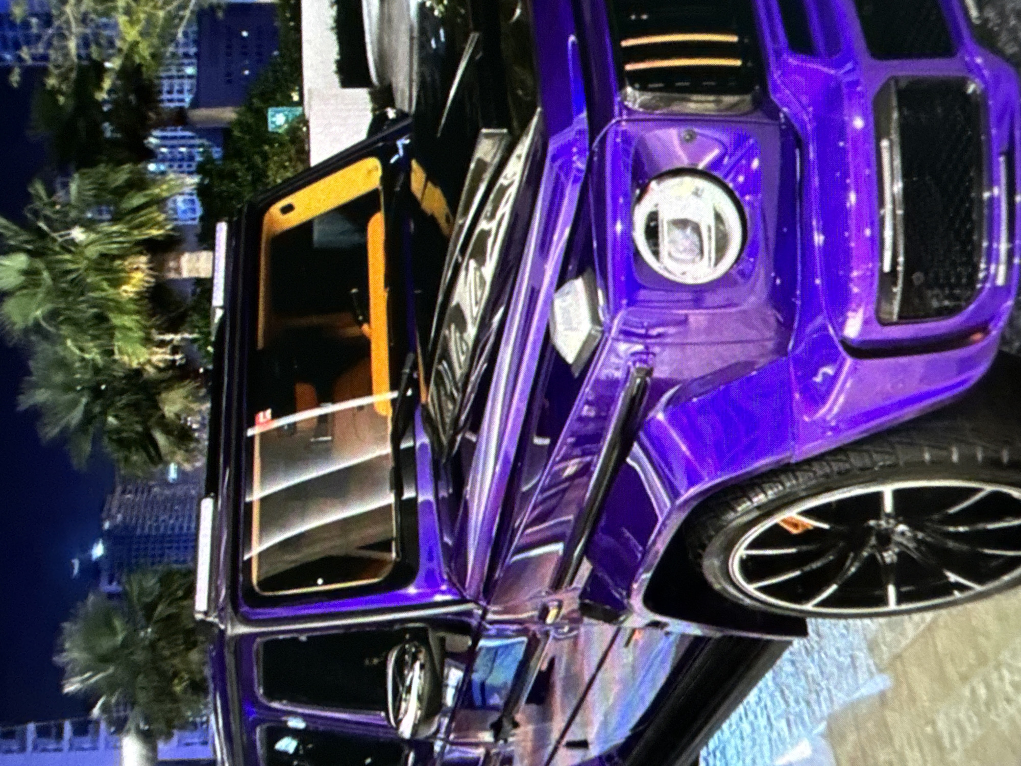 The image shows a purple Mercedes-Benz G-Class SUV parked outdoors, with its driver-side door open. The interior of the car is visible through the open door, with a tan or light brown color scheme. The vehicle's front tire and wheel are prominently displayed. In the background, palm trees and urban buildings, likely representing a cityscape at night, can be seen. The location is in the city of Unknown, Unknown.