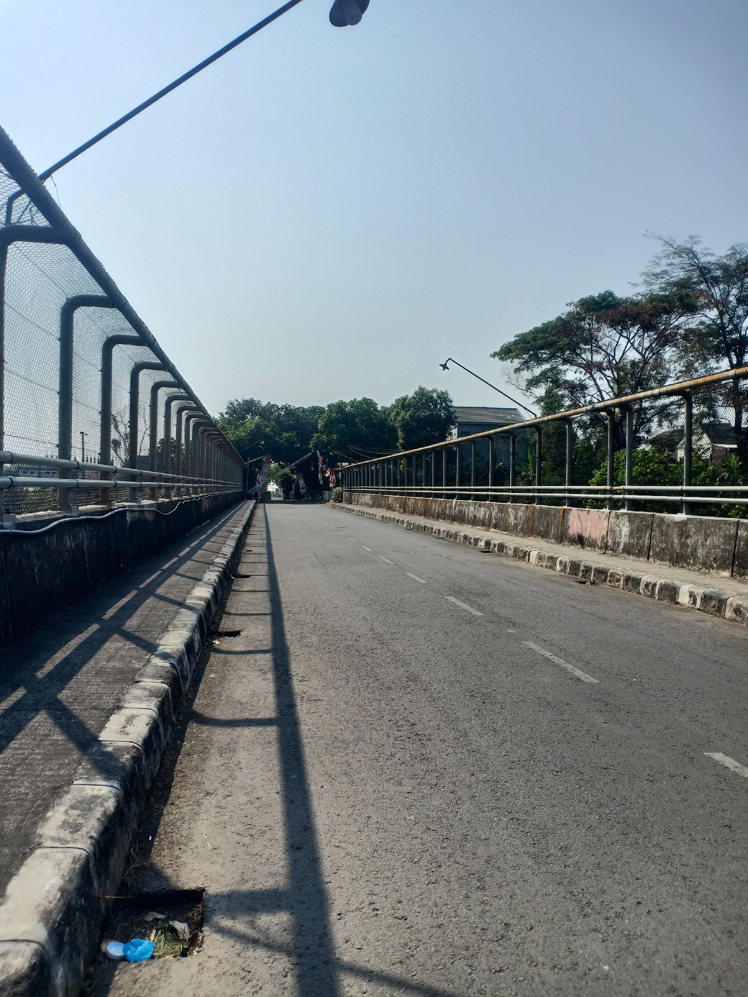 The image shows a view of a road on a bridge in the city of Semarang, Indonesia. The road is asphalt with white lines marking the lanes. On the left side of the road, there is a sidewalk with a concrete curb and a chain-link fence with metal poles. The fence casts shadows on the sidewalk and the road. On the right side of the road, there is a concrete barrier with a metal railing on top. Beyond the railing, there are trees and some buildings. The sky is clear and blue. There is some litter on the ground near the curb. In the distance, there are some people walking and a few flags are visible. The overall impression is of a sunny day on a somewhat aged bridge infrastructure.