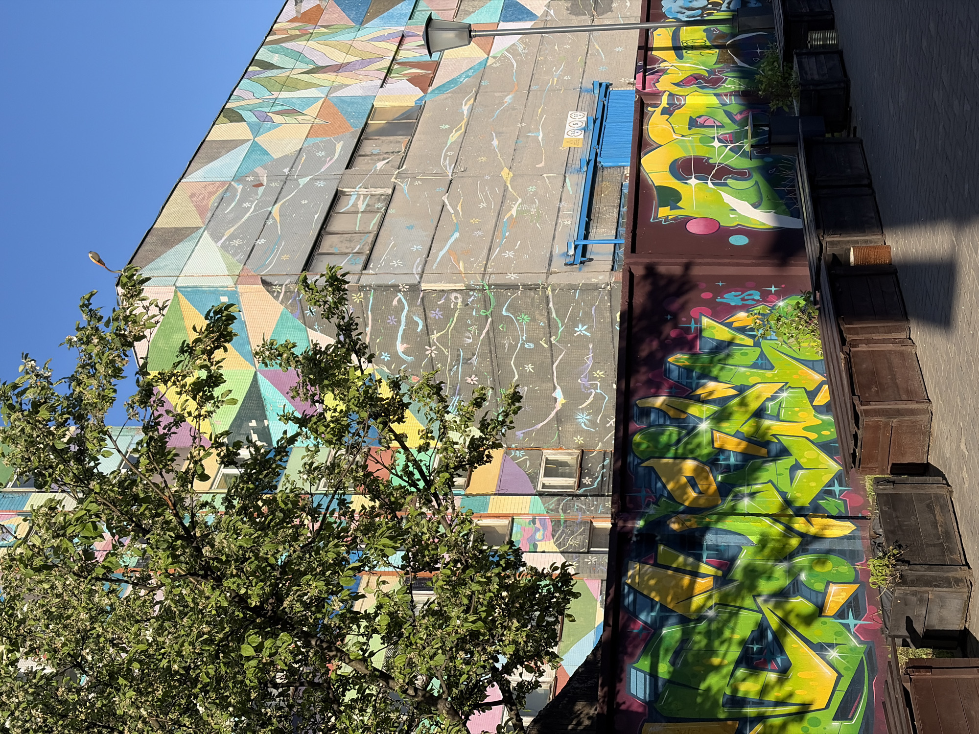 The image depicts the facade of a building in Minsk, Belarus. The building has two distinct sections: one with geometric, mosaic-style artwork, and another with graffiti. A tree with green foliage partially obscures the mosaic section. The graffiti section is painted in vibrant green and yellow, with darker maroon sections as well. Along the bottom edge of the graffiti section are a row of dark, rectangular planters. A street lamp is visible extending from the building. The sky is clear and blue. The image suggests a vibrant urban environment with street art and greenery.