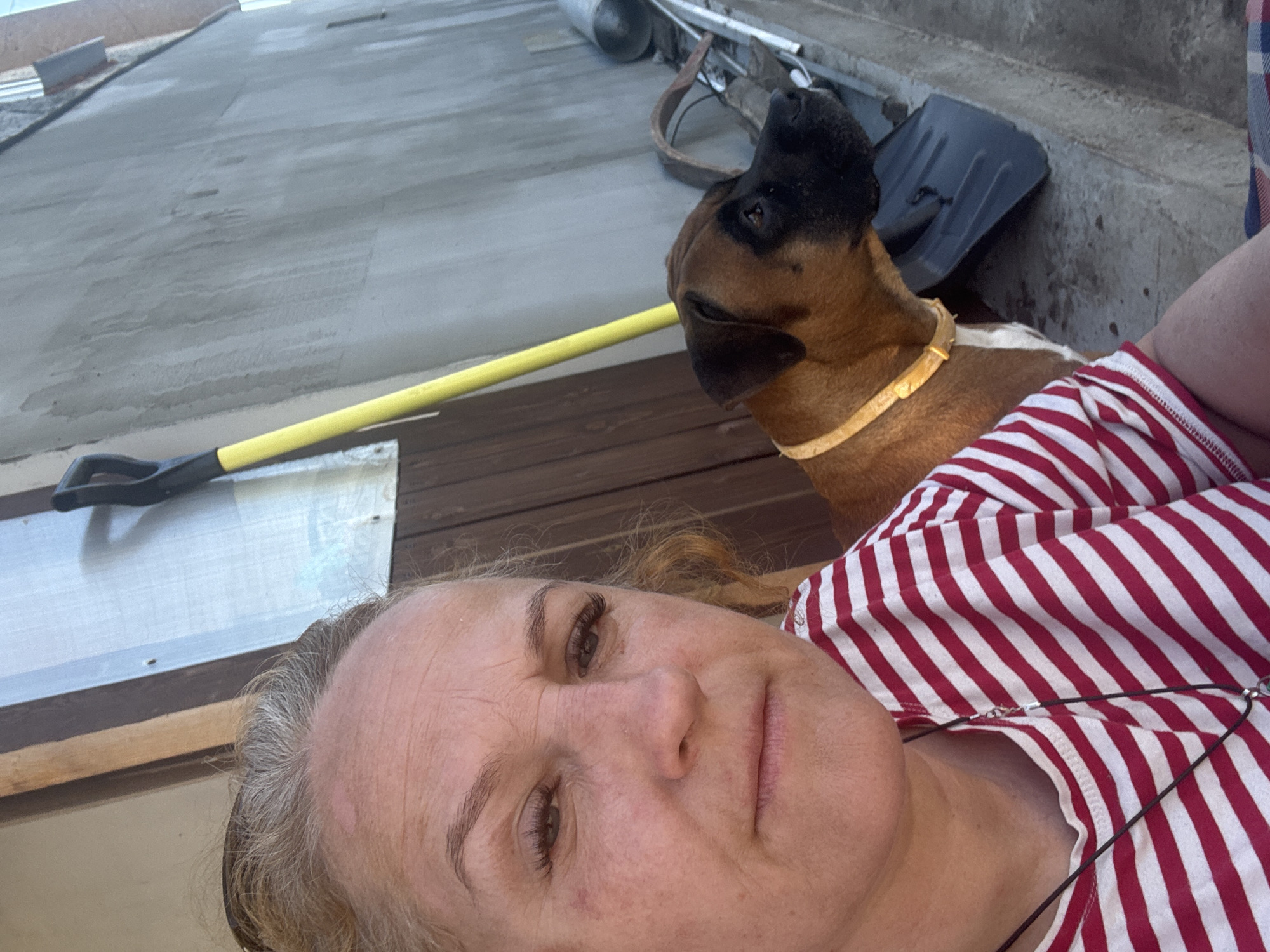 A middle-aged woman takes a selfie, partly obscuring the view of the environment. The woman has light skin, fine wrinkles, and light hair. She wears a red and white striped shirt and a necklace with a pendant. Beside her is a brown and black dog with a yellow collar. In the background, there is a gray concrete wall, a wooden deck, a yellow shovel leaning against the wall, and other miscellaneous objects. The location appears to be outdoors, possibly in a rural or suburban area. The city of Chaikovsky, Russia, cannot be verified from the visual information provided.