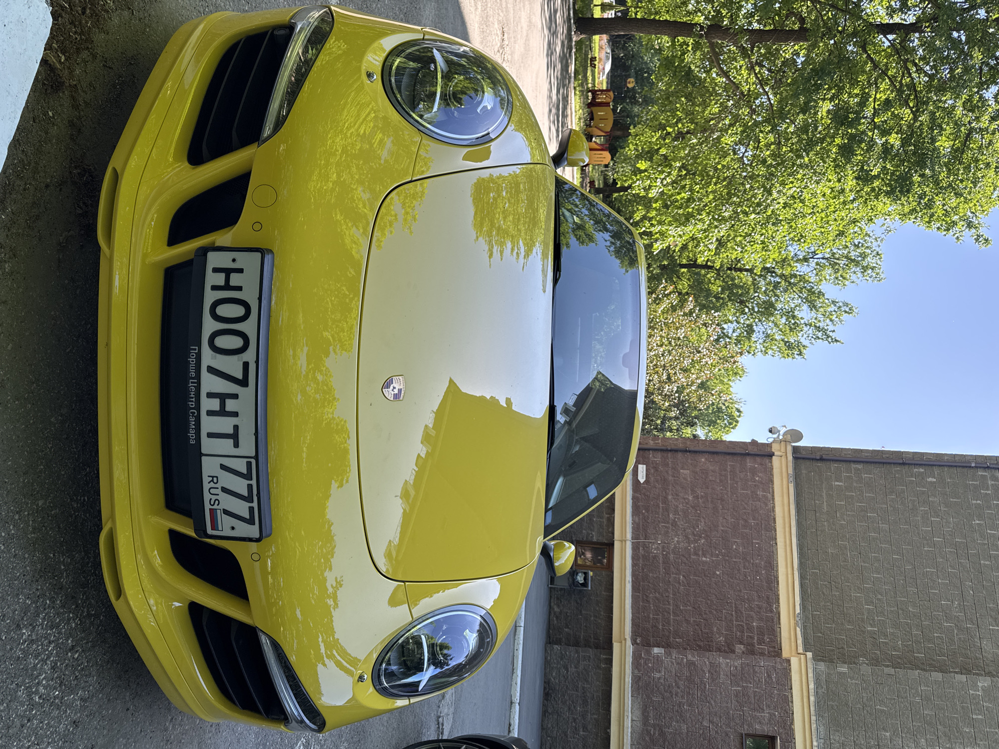 The image shows the front of a yellow Porsche parked on a paved surface, possibly a street or parking lot. The car has a Russian license plate reading "H007HT 777". The background includes a brick building, a tree, and a blue sky. The image is taken during daylight.