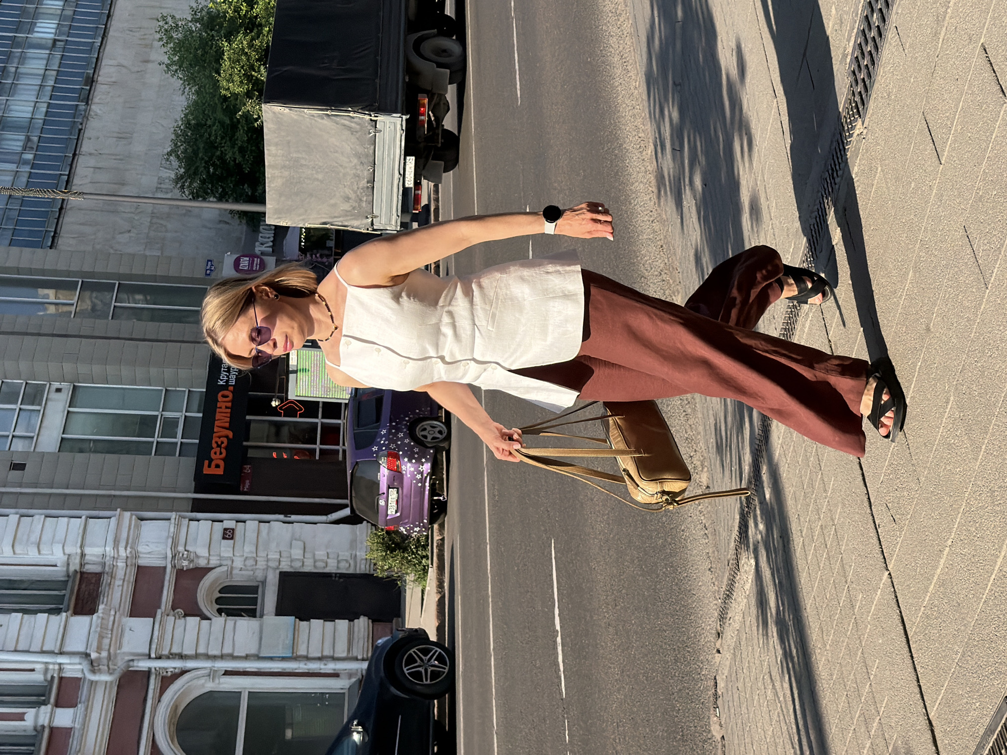 A woman stands on a sidewalk in Krasnoyarsk, Russia, bathed in the warm light of a sunny day. She is wearing a white sleeveless top and brown wide-leg pants, accessorized with black sandals and a light brown shoulder bag. A smartwatch is visible on her left wrist. Her blonde hair is styled in a bob, and she wears sunglasses. She is looking to her left, her gaze angled downwards. The background features a street with a truck, a purple car adorned with white stars, and buildings with varying architectural styles. One building displays an orange sign with Russian text. The scene appears to be set in an urban environment.