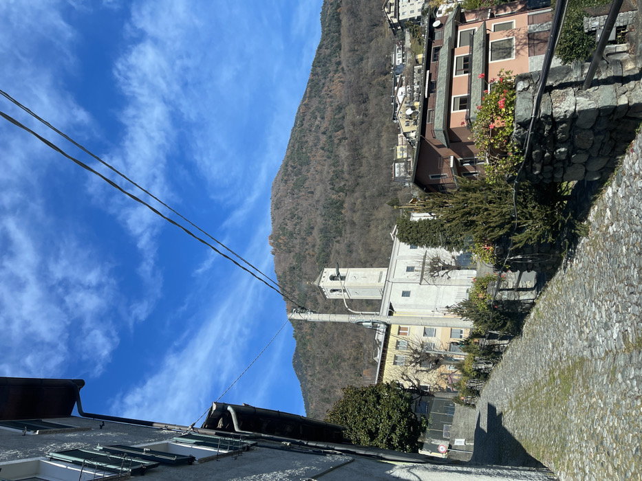 The image displays a picturesque mountain town, likely Castione, Italy, under a clear, bright blue sky adorned with wispy white clouds. The scene is viewed from an elevated perspective, looking down a sloping, cobblestone-paved street that curves gently to the right.
In the mid-ground, a prominent light-colored church with a tall, slender bell tower stands out. Surrounding the church are several multi-story residential buildings, showcasing traditional European architecture with a mix of roof styles, including dark tiles and terracotta hues. Many buildings feature windows with shutters and balconies, some adorned with planters holding vibrant red flowers, possibly geraniums.
A large, tree-covered mountain dominates the background, with the trees appearing mostly bare, suggesting a late autumn or winter season. Utility lines are visible traversing the sky above the town. In the immediate foreground, the edge of a building's roof and windows are seen on the left, while a rugged stone wall borders the cobblestone pathway on the right. Patches of evergreen foliage are interspersed among the buildings, providing green accents.
The lighting indicates a sunny day, casting clear shadows. No people are visible, and there is no discernible text on any of the structures or signs. The overall atmosphere is one of serene tranquility, characteristic of a quiet, traditional European village nestled in a mountainous landscape.