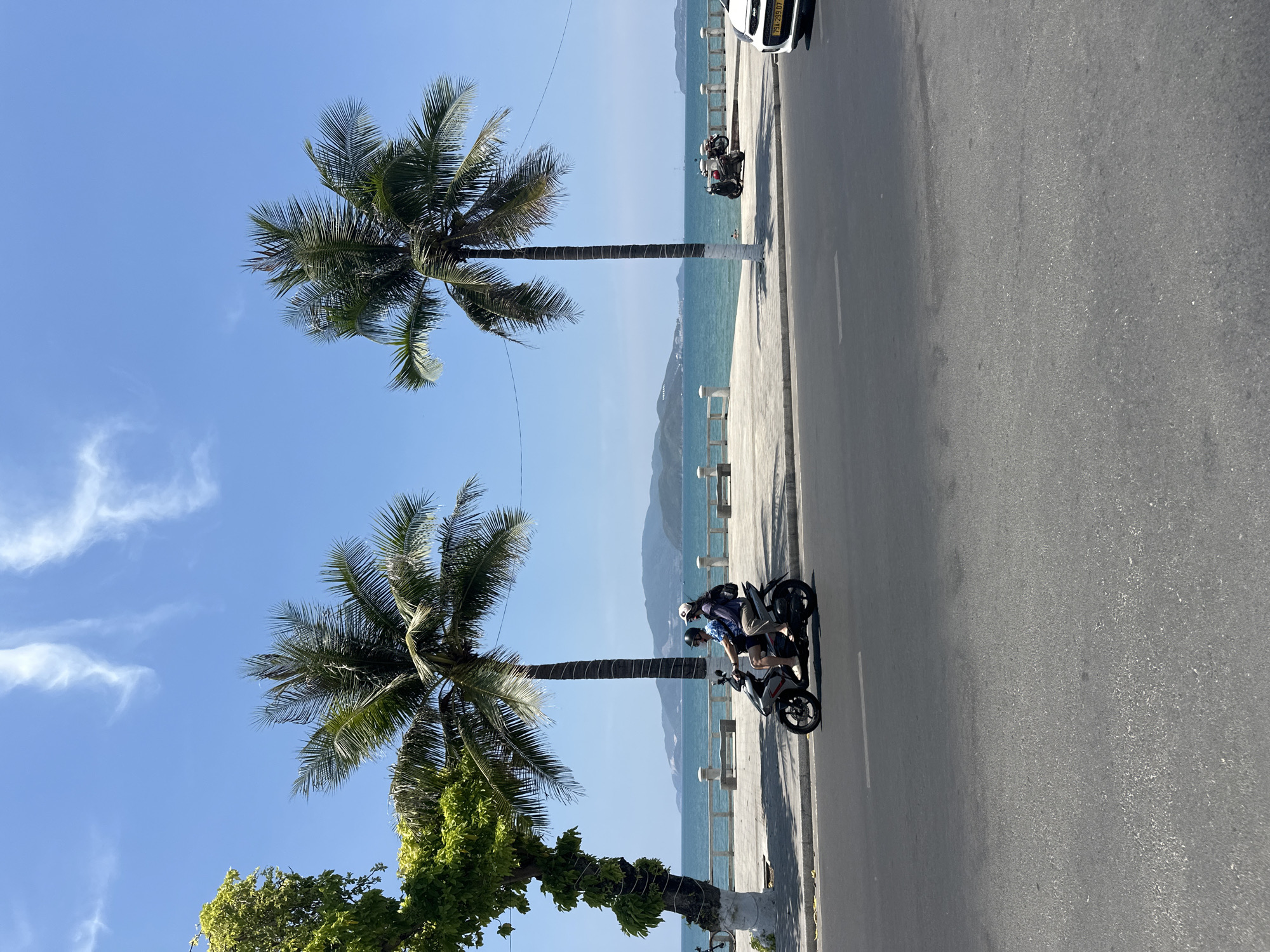 The image depicts a street scene in Nha Trang Ward, Vietnam. Two palm trees are prominent on the left, framing a view of the ocean and distant mountains. A paved road runs along the right side of the frame. Two motorbikes are visible on the road, one closer with two riders and another further away with one rider. A white car is partially visible in the top right corner of the image. The sky is blue with scattered clouds. The scene suggests a coastal environment with tropical vegetation.