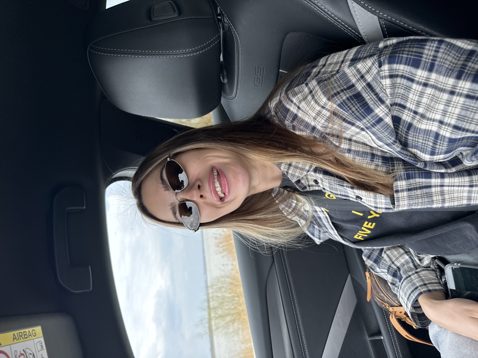 The image captures a female individual seated in the passenger side of a vehicle located in Pasohlávky, Czechia. The subject, with long, light brown hair and blonde highlights, is looking directly at the camera with an open-mouthed expression and her tongue slightly extended. She is wearing small, oval-shaped sunglasses with dark lenses and a thin metal frame. Her attire consists of a dark t-shirt with partially visible yellow text reading "FIVE YEA" layered under an open, long-sleeved flannel shirt featuring a blue, dark blue, and off-white plaid pattern. A patterned brown purse is visible on her lap, and her left hand holds a dark-colored smartphone. The vehicle's interior includes black leather seats with white contrast stitching, displaying a "GS" logo on the passenger seat's headrest. An "AIRBAG" warning label is affixed to the beige sun visor above. Through the passenger window, an overcast sky hangs above a wide body of water, likely a lake or reservoir, with a visible strip of light brown vegetation along its distant shore, characteristic of the Pasohlávky region.
