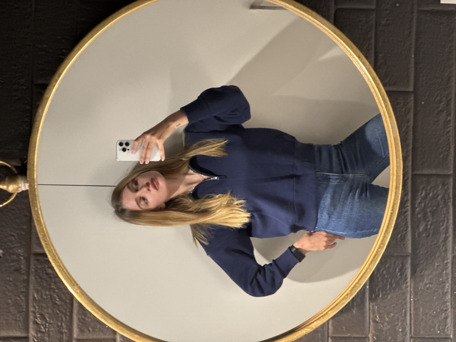 A young woman with long blonde hair, wearing a navy blue quarter-zip sweatshirt and blue jeans, is captured in a mirror selfie taken indoors in Tbilisi, Georgia. She holds a white smartphone with multiple rear cameras in her raised right hand, aiming it towards the mirror, while her left hand rests on her hip. A dark-colored smartwatch is visible on her left wrist. Her gaze is directed slightly to the right, away from the camera. The reflection is framed by a circular mirror with a prominent gold-colored ornate frame. Behind the woman, a plain white wall is visible within the mirror's reflection. The wall surrounding the mirror is clad in dark, rectangular, possibly brick-like, textured tiles.