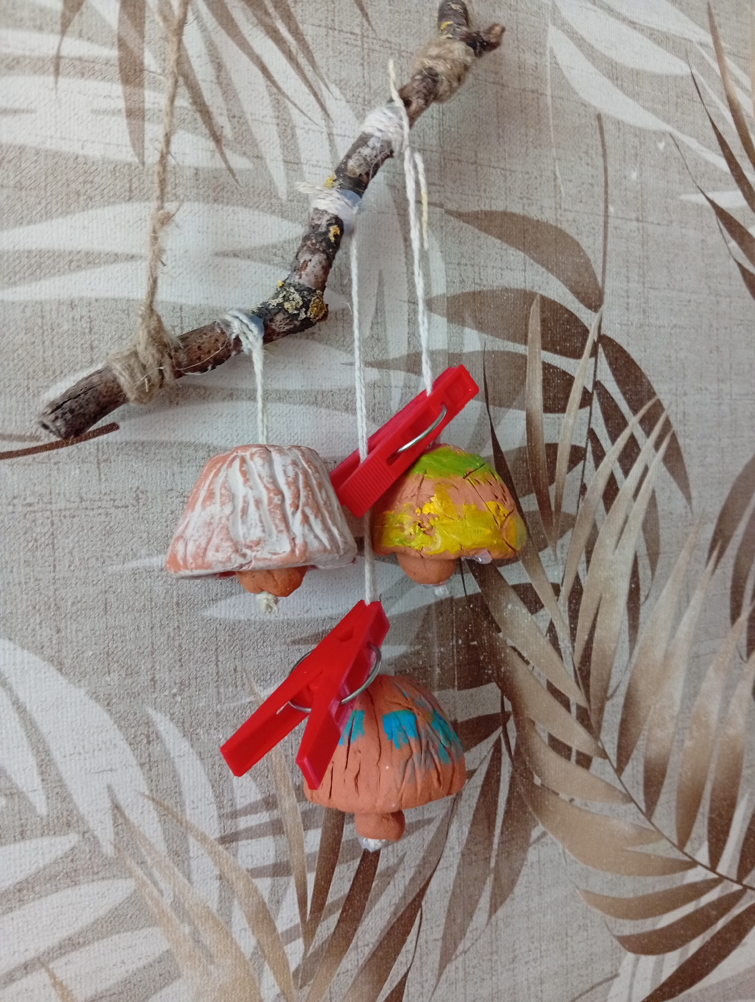 The image shows a handmade mobile hanging on a wall with a leaf-patterned wallpaper. The mobile consists of a rough-textured twig from which three small, bell-shaped objects are suspended by white strings. These objects appear to be made of clay and resemble mushrooms. The first mushroom from the left is primarily orange with white detailing. The middle mushroom is painted with green and yellow colors. The mushroom on the right is orange with splashes of blue paint. Two red plastic clothespins are attached to the strings of the middle and bottom mushrooms, likely used to hold them in place or for decoration. A rough twine is wrapped around the twig on the left side of the mobile. The lighting suggests an indoor setting. There are no people or animals visible in the image. The location is not identifiable from the image itself and the specific mention of Baranavichy, Belarus, cannot be confirmed.