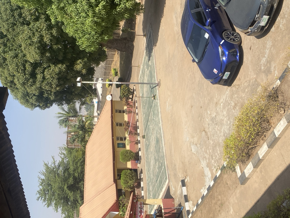 This image depicts an outdoor compound, likely a school or recreational facility, located in Tudun Nupawa, Nigeria, under bright daylight conditions. The scene is viewed from an elevated position, with a roof overhang visible at the top left.
In the mid-ground, a single-story building with yellowish walls and a red-brown corrugated metal roof extends horizontally. It features multiple rectangular windows and is fronted by landscaped shrubs. To its left, a partial view of another structure with a similar roof color displays a sign partially legible as "N-CHICKENS."
Immediately in front of the main building, a rectangular court, painted light green with white boundary lines, is visible. A net is strung across its center, indicative of a badminton or volleyball court. A tall, white metal pole equipped with floodlights stands adjacent to the court on the right. Further right, a satellite dish is mounted on a pole near the building.
The ground surrounding the court and structures is primarily unpaved dirt or a worn concrete surface. Black and white painted curbs define edges of pathways and a small, shrub-filled planter in the foreground.
Two sedans are parked in the lower right quadrant. The foremost vehicle is a dark blue Toyota Corolla, displaying an "L" plate (learner's permit) on its front bumper and windshield. Its license plate originates from "KANO." Partially obscured behind it is a darker-colored sedan, also with a "KANO" license plate.
The background features numerous green trees, including a very large specimen on the right. A raised, rectangular structure, possibly a water tank, is visible among the trees further back. A block wall delineates the property perimeter in the distance. No individuals are visible in the scene.