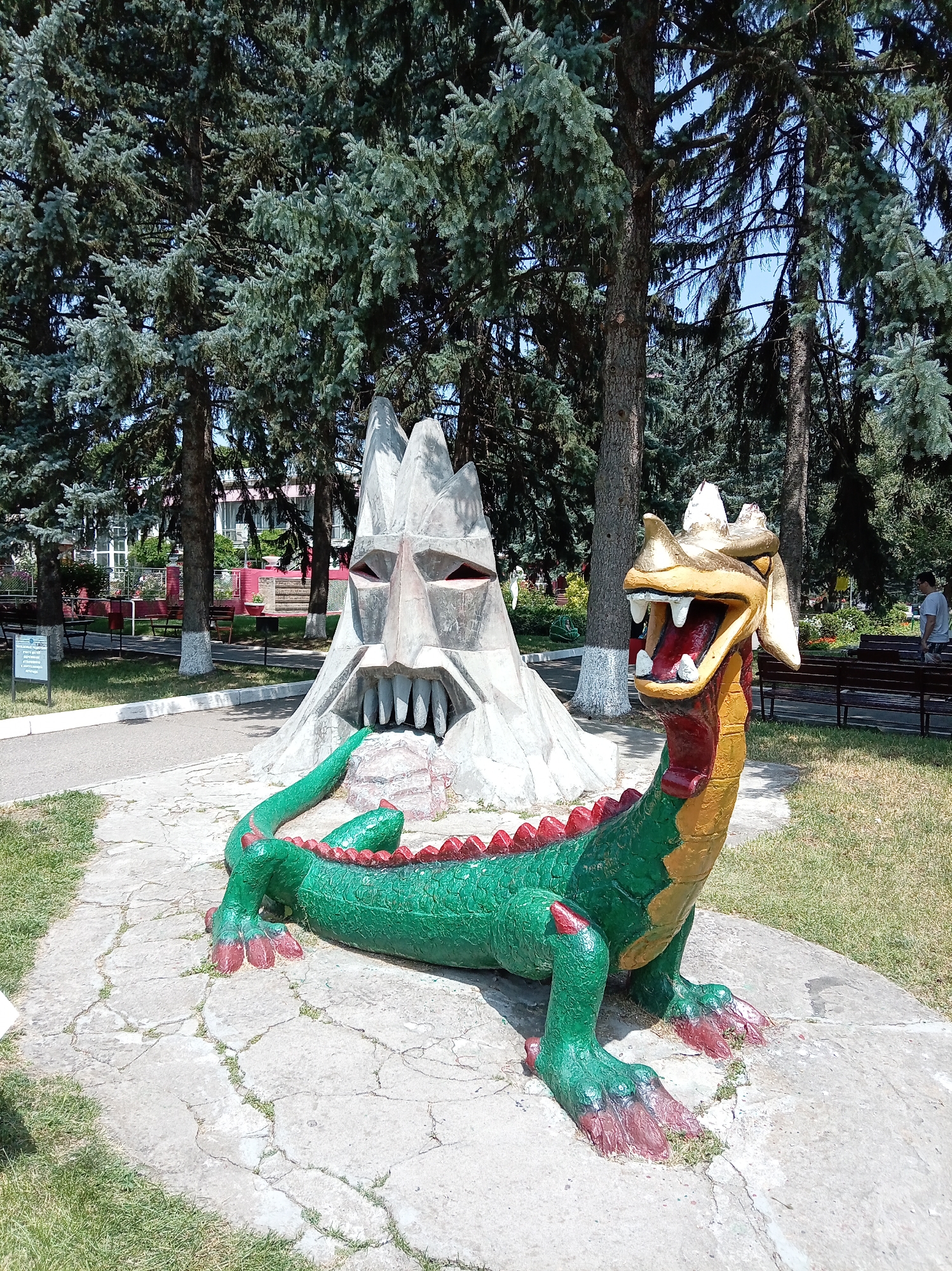 This image depicts a park area with concrete statues and lush green trees. In the foreground, a brightly colored statue of a green dragon with red spikes and yellow accents is positioned on a concrete base. To the left of the dragon, a large, abstract concrete sculpture with a face-like appearance and sharp teeth is visible. The background shows more trees, some benches, and a building. A young man in a light-colored shirt is walking in the distance on the right. The scene appears to be from a public park in Yessentuki, Russia, likely within the city's recreational areas given the presence of decorative statues and well-maintained grounds.