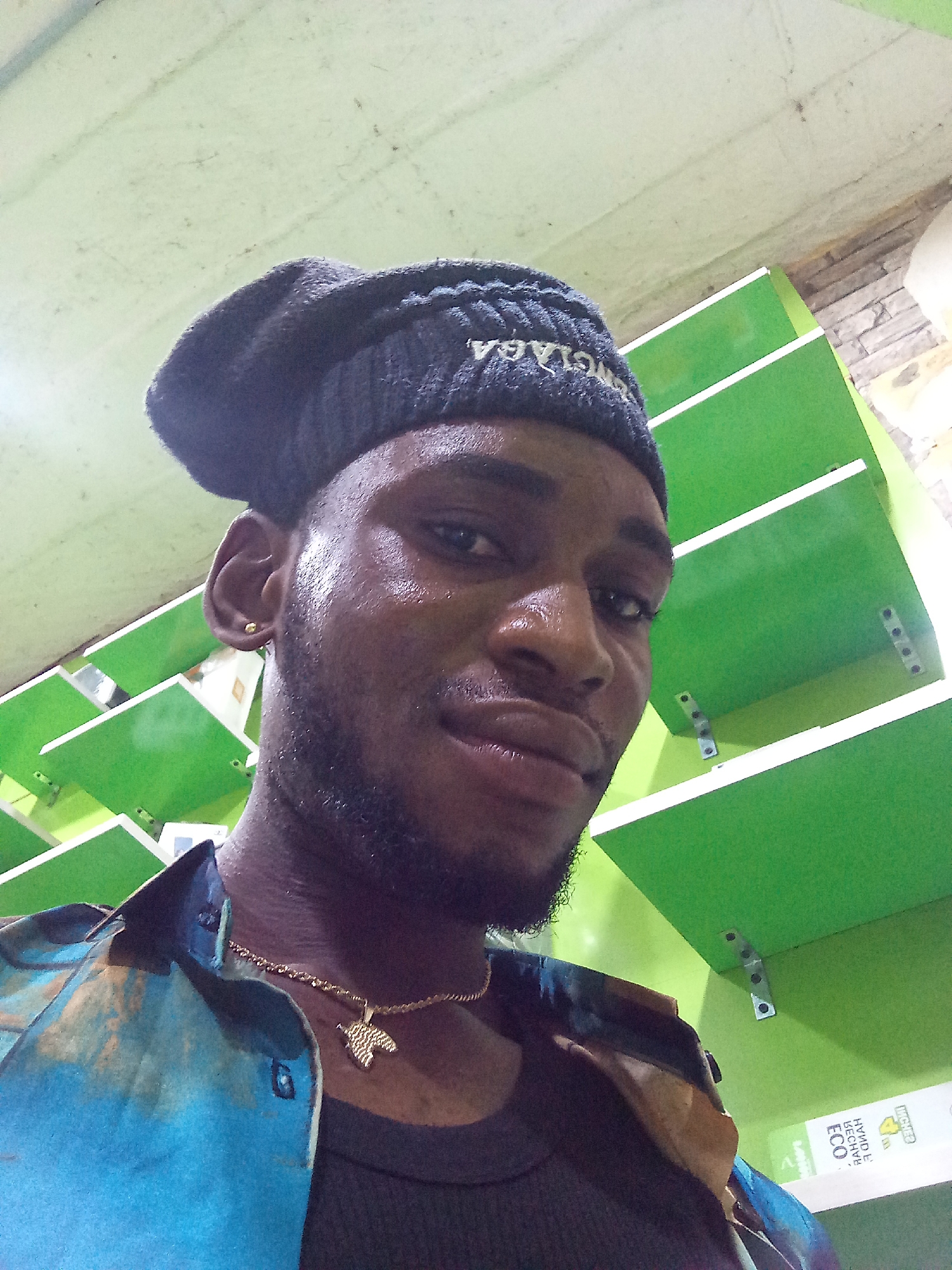 The image is a selfie taken from a low angle, showing a young Black man wearing a dark beanie with white lettering, a gold chain with a leaf-shaped pendant, and a colorful patterned shirt over a dark undershirt. He has a light beard and a gold stud earring in his left ear. The background consists of green shelves against a pale green ceiling and a tiled wall on the right. The lighting suggests an indoor setting, possibly a store or market, common in Lokoja, Nigeria. The man is looking directly at the camera, and his skin appears to be slightly sweaty, indicating the climate or exertion.