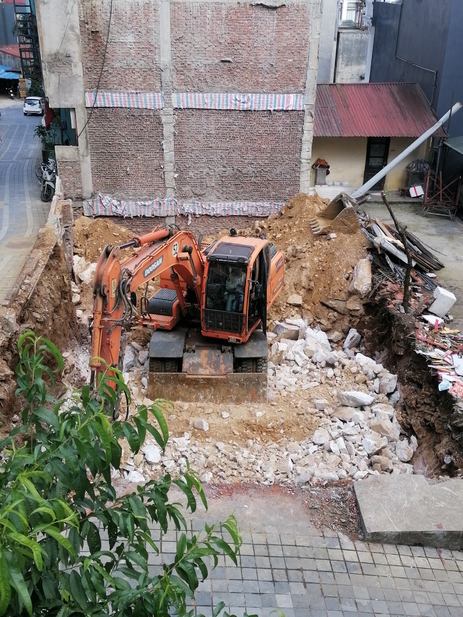 The image shows an excavation site in Sa Pa Ward, Vietnam. An orange Doosan excavator is the central subject, positioned within a pit filled with rubble and soil. The excavation is bordered by brick buildings, one with striped material draped across it. Debris and construction materials are scattered around the perimeter of the pit. A small red-roofed structure is visible in the background. In the foreground, green foliage obscures part of the scene. The area around the excavation is paved with gray square stones. In the distance, a vehicle is visible on a street.